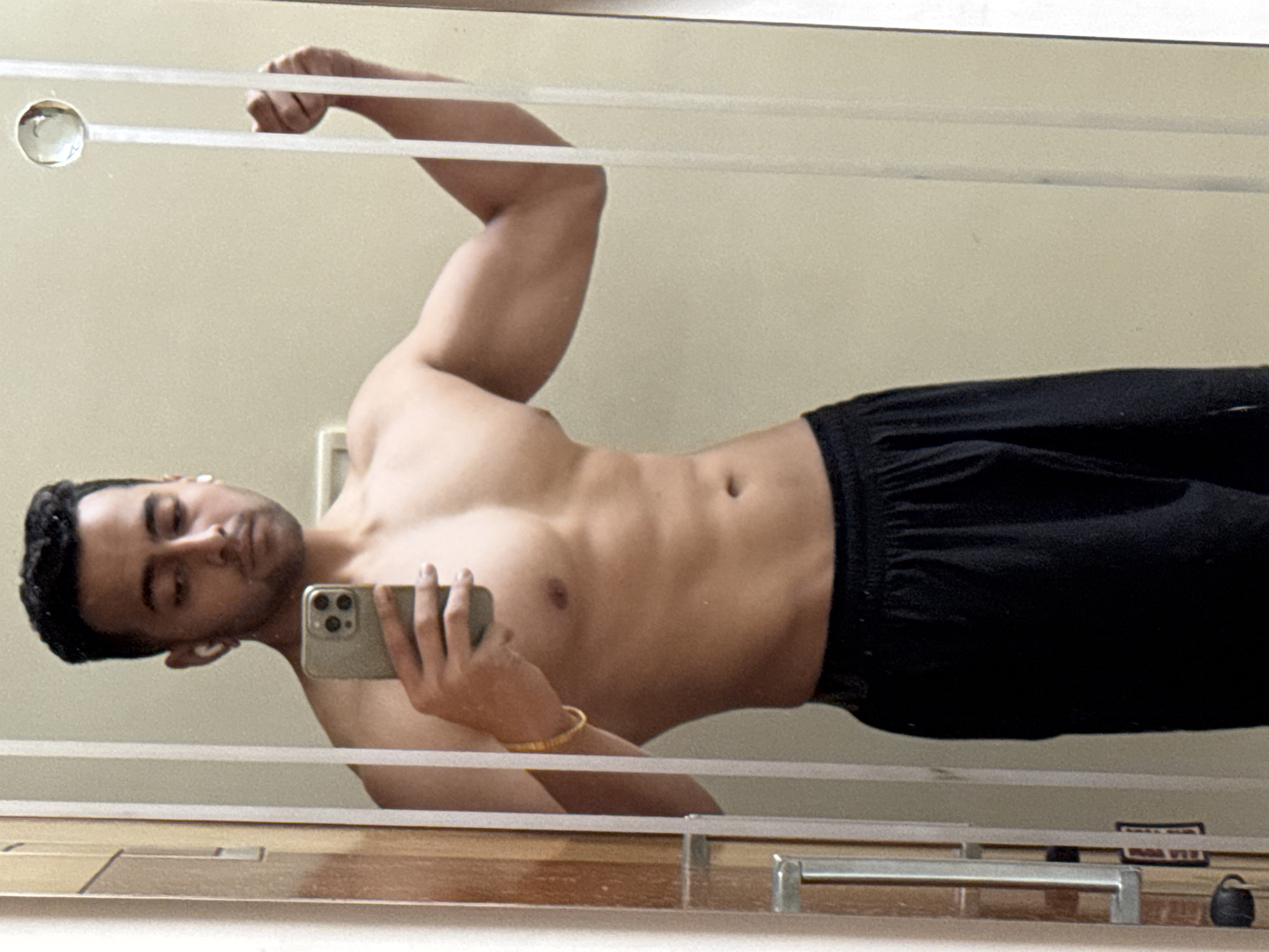A man is taking a selfie in a mirror. The man is flexing his right bicep. He is shirtless and wearing black shorts. He is holding a smartphone in his left hand. He is wearing a bracelet on his left wrist. He has dark hair and a light complexion. He is wearing wireless earbuds. The background is a plain wall and a wooden frame. The scene is indoors. There is no evidence to suggest the photo was taken in Kolkata, India.