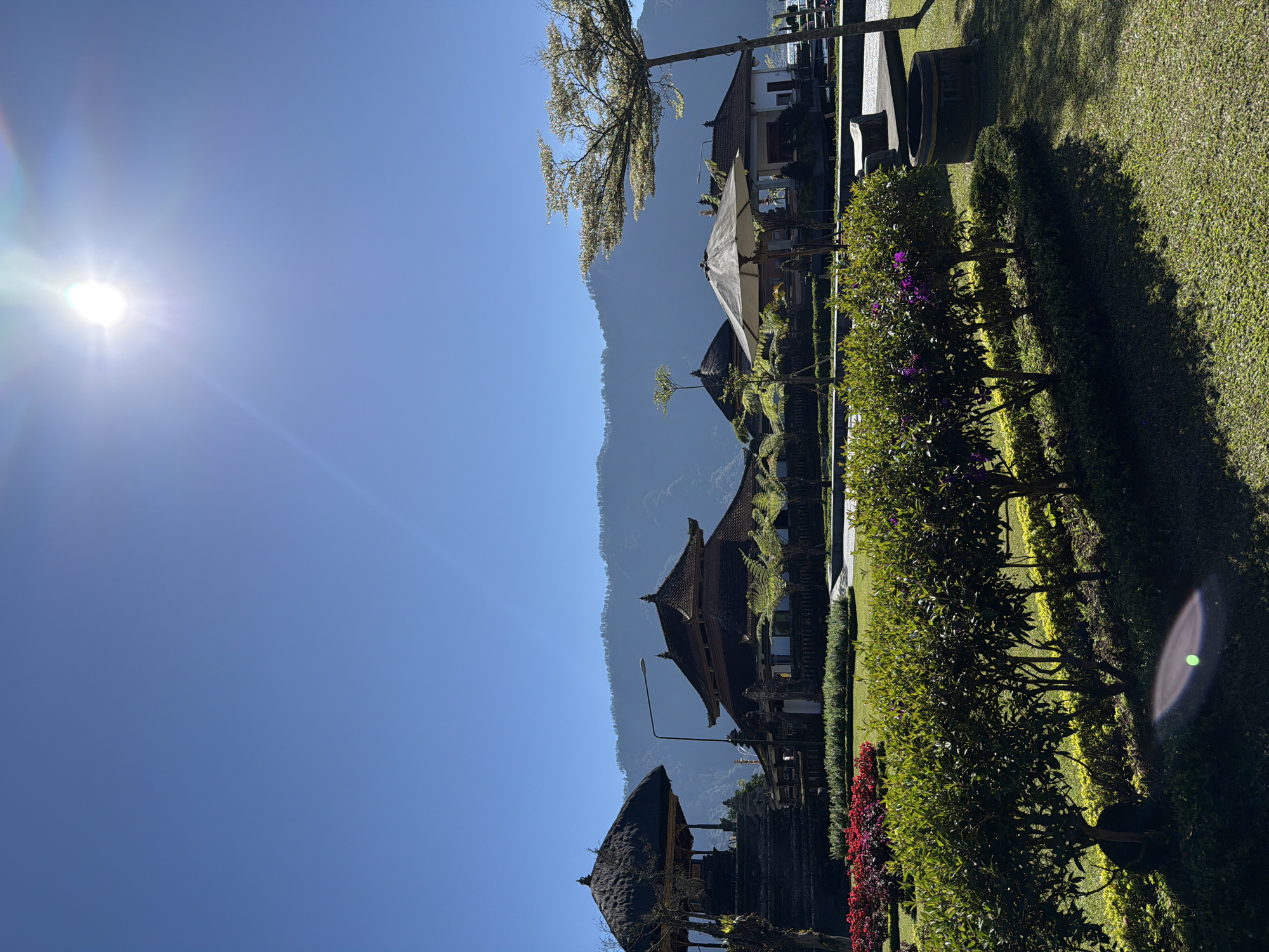 The image captures a view of Pura Ulun Danu Bratan, a water temple complex located in Baturiti, Bali, Indonesia, on a bright, sunny day. The sky is a clear, vibrant blue with the sun prominently positioned in the upper right quadrant, casting a bright glare and lens flare. In the foreground, a meticulously maintained garden features neatly trimmed green bushes and a strip of red flowers. Beyond the garden, several traditional Balinese structures with tiered, dark-tiled roofs are visible. One structure on the left has a thatched roof. A large white umbrella provides shade near one of the buildings. In the background, hazy, verdant mountains rise against the horizon. The overall scene conveys a sense of tranquility and cultural beauty.