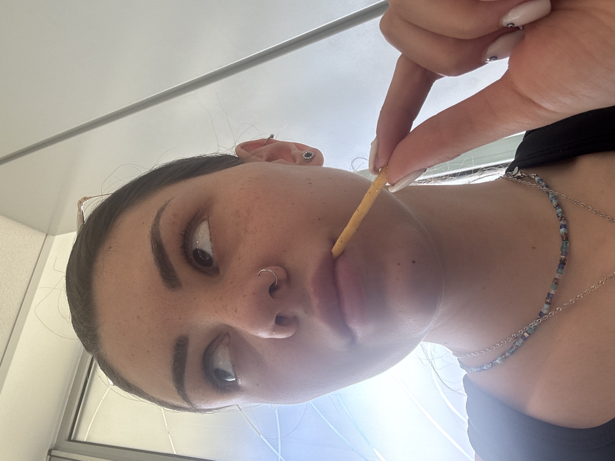 A close-up, low-angle portrait captures a young person, likely female, looking directly upwards towards the camera. Her head is tilted, creating an engaging and intense gaze. She is shown in the act of bringing a long, thin, light-colored stick-like snack (possibly a breadstick) to her mouth, held by her right hand.
She has dark, well-defined eyebrows, light brown eyes, and freckles visible across her nose and cheeks. A silver hoop nose ring adorns her left nostril. Her left ear displays a dark circular stud earring and a small silver ring in the helix. Part of a pair of eyeglasses is visible, resting on her hair or forehead.
She wears two layered necklaces: one a strand of small, colorful square beads in shades of blue, green, and brown, and another delicate silver chain. Her attire appears to be a dark-colored top. Her right hand features long, light-colored fingernails, some embellished with a small blue circular design.
The background is uniformly light and out of focus, suggesting an indoor setting with a high ceiling or wall. Bright light emanates from above and behind the subject, casting a soft glow and creating some glare. There is no visible text. The image does not contain specific visual cues to confirm the location.