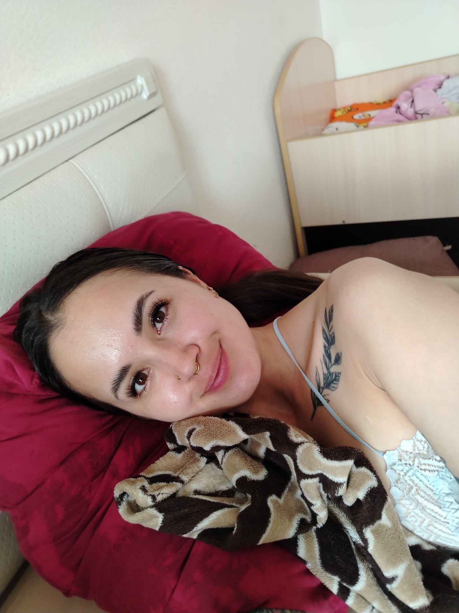 A young woman with dark hair and brown eyes is lying on a bed with a red pillow. She is wearing a light blue top with lace trim and has a tattoo of a branch with leaves on her left shoulder. A brown and beige patterned blanket is draped over her. In the background, a wooden crib filled with blankets and pillows can be seen. The scene appears to be a bedroom, possibly in Noviy, Russia.