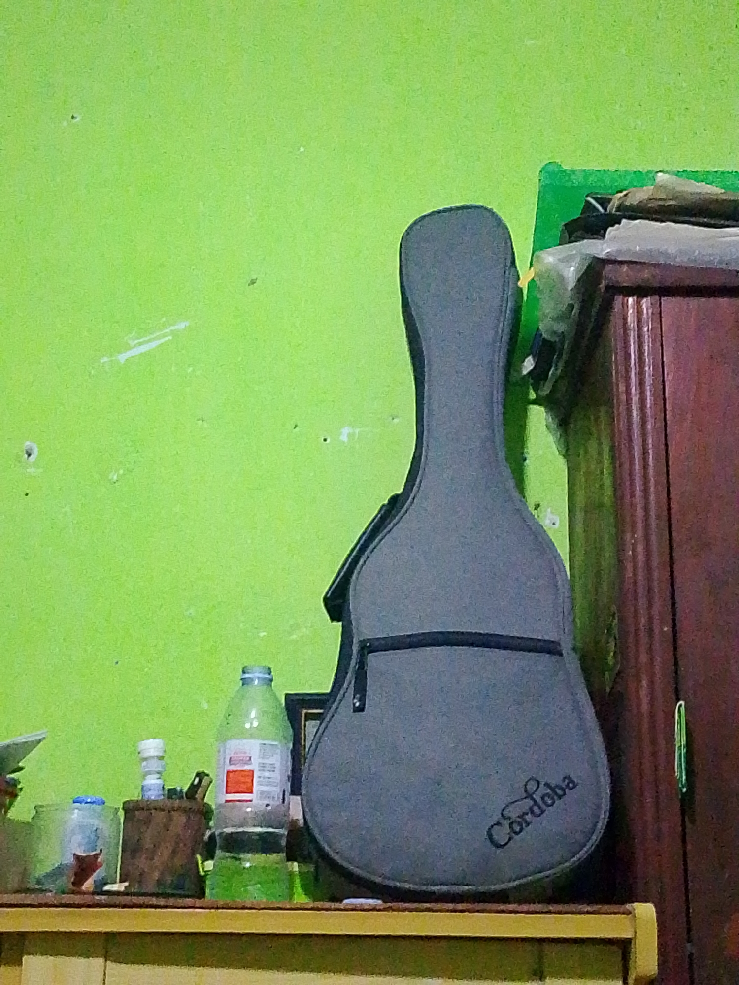 A gray guitar case with the word "Cordoba" embroidered in script on its lower bout stands upright against a lime green wall. To the left of the guitar case, on a wooden shelf, are two plastic water bottles, one partially filled with clear liquid and the other with a green liquid, and a small woven basket. To the right of the guitar case is a dark brown wooden cabinet, partially obscured by the guitar case. The cabinet has vertical grooved detailing and a plastic bag containing various items rests on top of it. The scene is brightly lit, suggesting an indoor setting. The overall atmosphere is simple and utilitarian. The image appears to be taken in Banyuwangi, Indonesia.