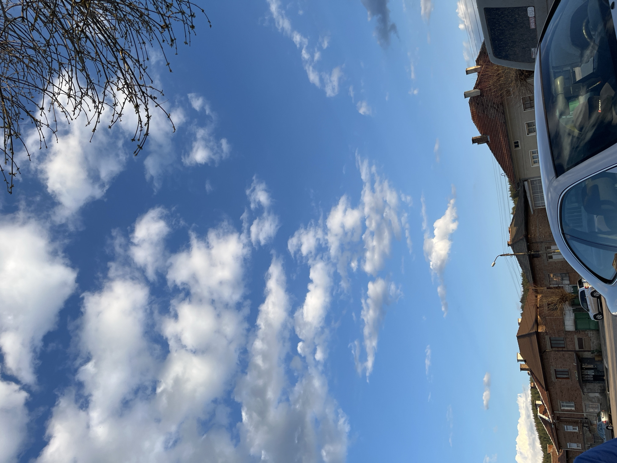 The image shows a view of Karavelovo, Bulgaria, with a blue sky filled with scattered white clouds dominating the scene. In the upper left corner, bare tree branches are visible. On the right side, residential buildings with red tile roofs can be seen, and part of two cars are visible and parked on the right side of the frame. A street lamp is also visible near the buildings.