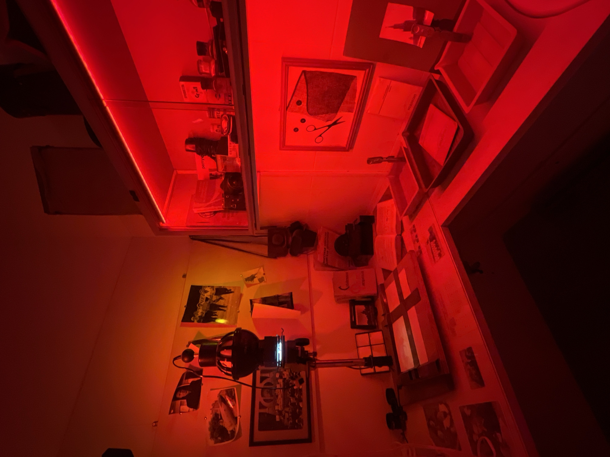 The image shows a darkroom lit by a red safelight. A photographic enlarger is visible on the left, with prints and negatives pinned to the wall around it. Shelves above the table are stocked with bottles and containers. Trays for developing chemicals are on the table to the right, alongside a framed artwork of a pair of scissors cutting a piece of fabric. The location is unknown and there is no indication that it is in Saint Petersburg, Russia.