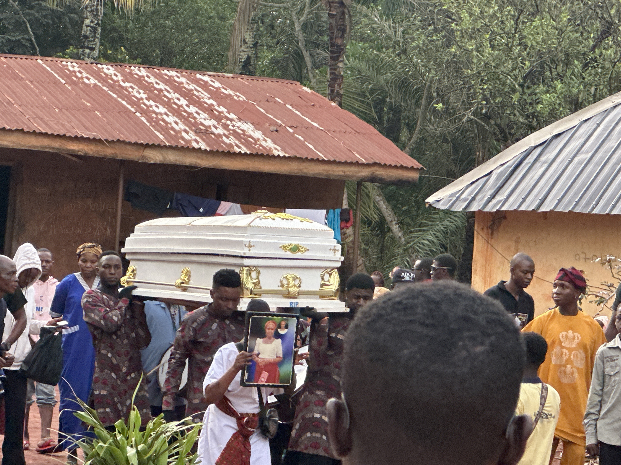 In an outdoor setting in what is suggested to be a rural location, a funeral procession is underway. Six men are carrying a white coffin adorned with gold-colored details. Ahead of them, a person in white attire is holding a framed photograph of the deceased. Various individuals, dressed in a range of clothing, follow the procession. The surrounding environment includes buildings with corrugated metal roofs and dense foliage.