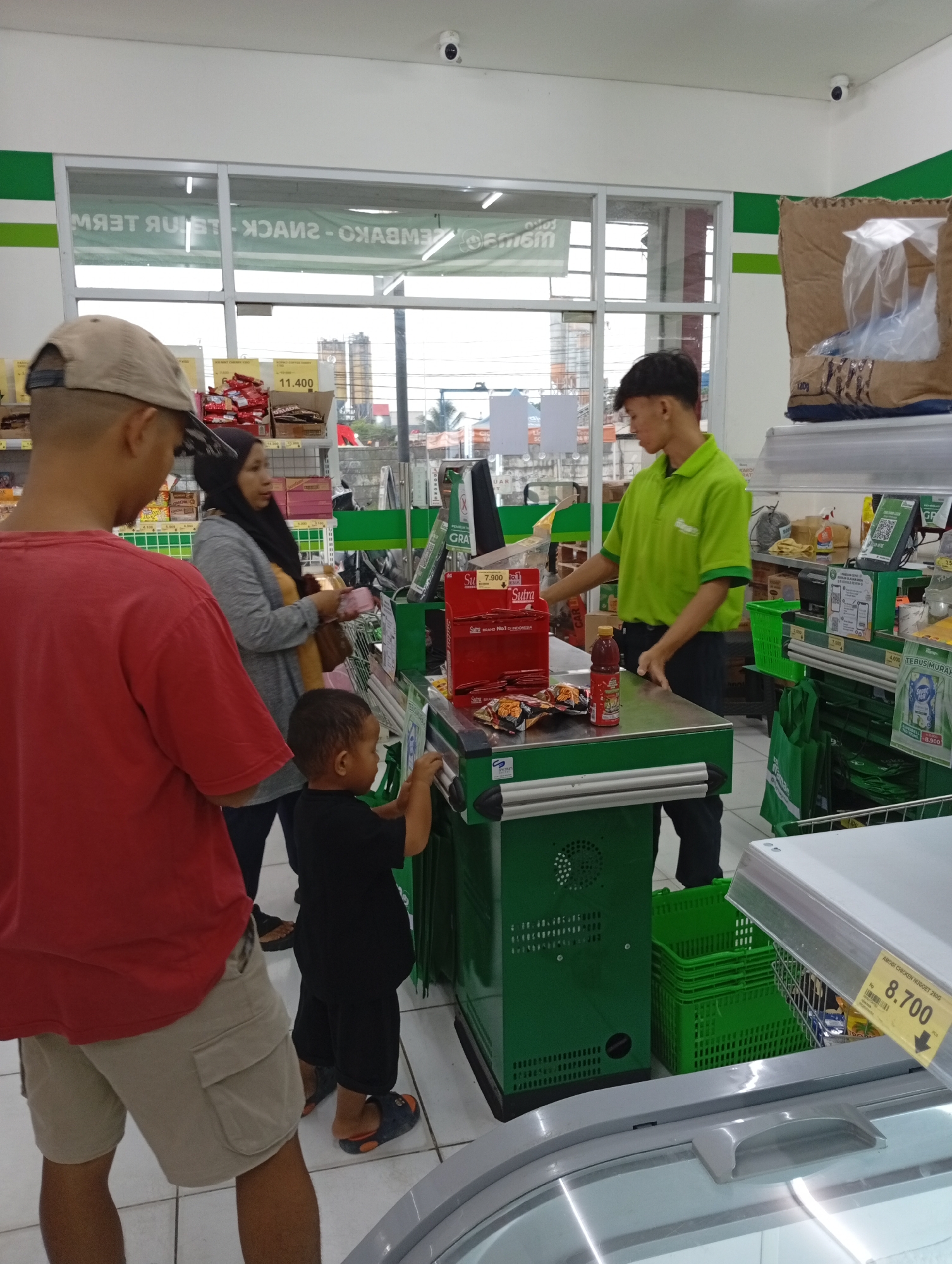 In a supermarket in Babakan, Indonesia, a cashier in a bright green uniform scans items at a checkout counter. A family stands in line, including a man wearing a red t-shirt and beige shorts, a woman in a grey hijab and sweater, and a young child in a black t-shirt. The child is reaching towards the counter, seemingly interacting with the scanner. The environment is a typical retail space with shelves stocked with products in the background and a refrigerated display case in the foreground. A sign displaying "AMOBY CHICKEN NUGGET 255G" and "Rp 8.700" is visible on the refrigerated case. The overall scene captures a mundane moment of everyday shopping.