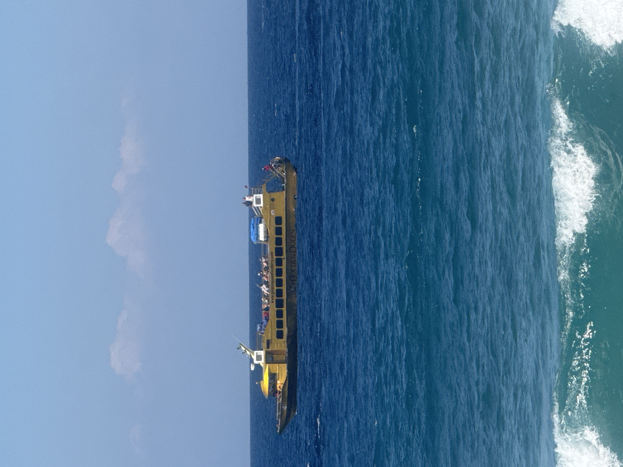 A yellow tourist boat, identified as "GOLDEN QUEEN," sails on a calm, deep blue ocean under a clear sky with scattered white clouds. The boat, with its windows, a small cabin, and a railing, is carrying several passengers who are visible on the upper deck. The boat's name is clearly printed on its side. The scene captures a serene moment of sea travel on a bright day, with the vastness of the ocean stretching out to the horizon.