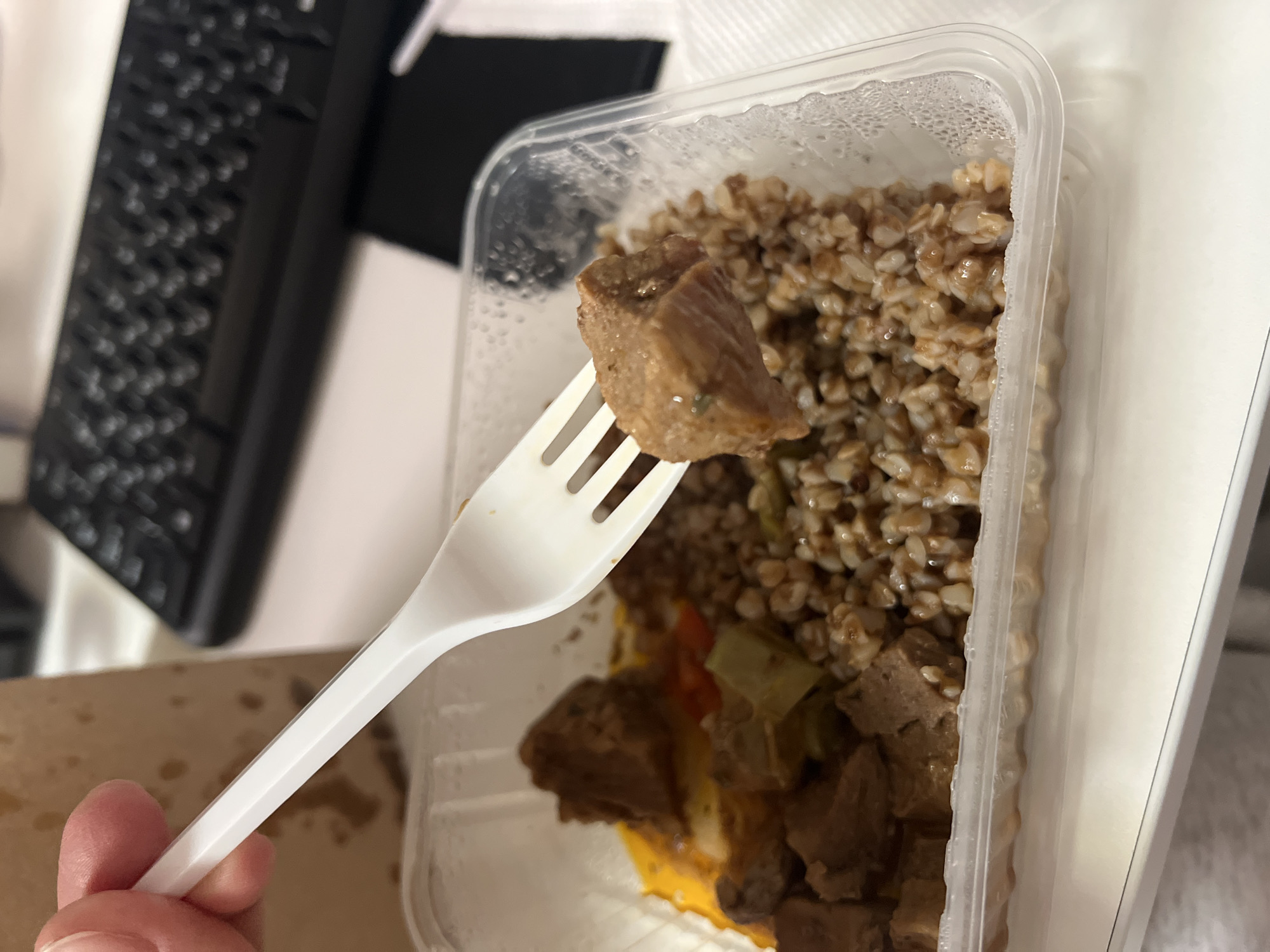 The image shows a partially eaten meal in a clear plastic container. The meal appears to consist of buckwheat and diced meat. A piece of meat is skewered on a white plastic fork held in a hand. In the background, a black keyboard is visible. The location is likely indoors, possibly an office or home in Khimki, Russia, given the user's request.
