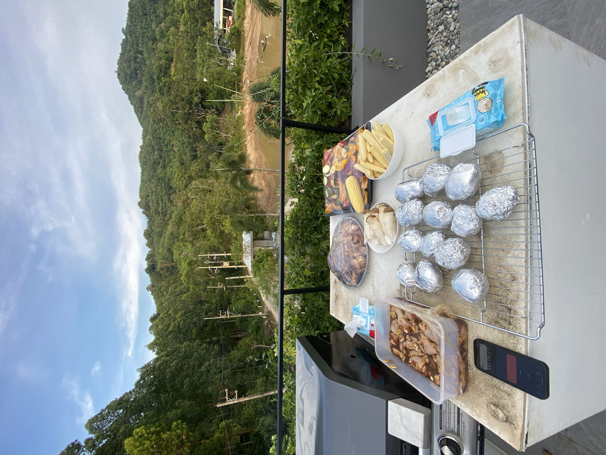 Here is a detailed description of the image:
**Scene:**
The image depicts a outdoor grilling setup on a patio or balcony, with a scenic view of a lush, hilly landscape. The location is likely in Choeng Thale, Thailand, given the presence of vegetation typical of the region.
**Objects:**
* **Grilling Station:** A grill is visible, with a gray table surface. Various food items are prepared for grilling.
* **Food:** There are several types of food ready for grilling. A container of corn, various vegetables, a platter of meat, and mushrooms can be identified. Additionally, there is a container of chicken, and potatoes wrapped in aluminum foil on a cooling rack.
* **Accessories:** A pack of baby wipes and a smartphone are also placed on the surface next to the grill.
* **Environment:** The scene is outdoors, with a view of a densely forested hillside and a body of water. Electrical lines are visible.
**Environment:**
* **Setting:** A balcony or patio with a lush, natural environment.
* **Weather:** The weather appears to be sunny.
**Summary:**
The image captures a cooking scene on a balcony or patio in a natural setting, likely Choeng Thale, Thailand. Various food items are prepared for grilling.