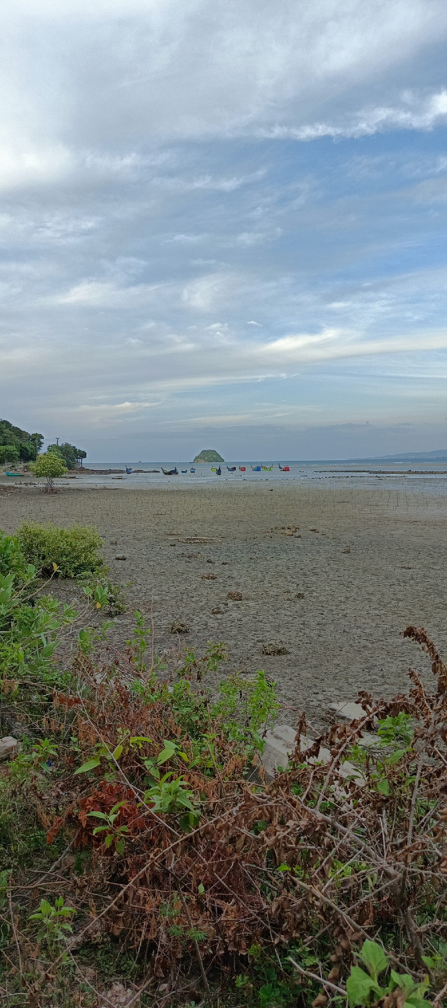 The image depicts a beach scene in Lampageu, Indonesia, during what appears to be low tide. The foreground is dominated by dry, cracked mudflats with scattered small rocks and vegetation. In the middle ground, a shallow body of water is visible, dotted with numerous small, colorful fishing boats anchored offshore. A small, tree-covered island is prominent in the distance, situated on the horizon. The background consists of a hazy sky with scattered clouds. The overall impression is one of a calm, natural coastal environment.