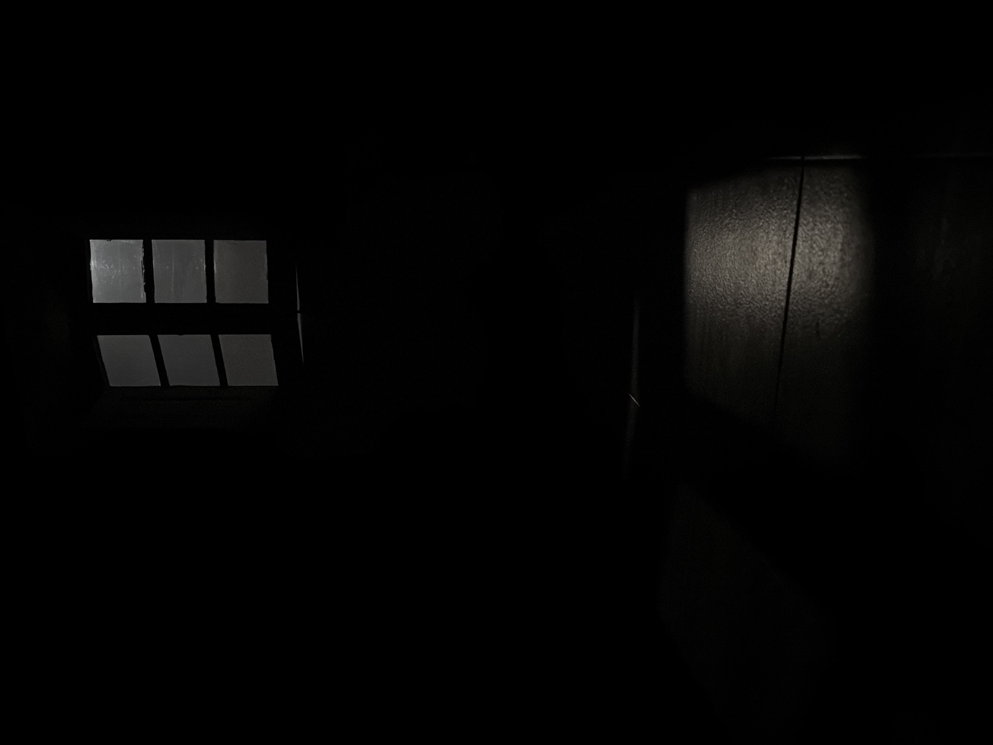 This media file depicts an extremely dark interior space, possibly a cellar or an old, dimly lit room in Sant Martí Sarroca, Spain. No people or specific activities are visible.
On the left side of the frame, a rectangular window is the primary source of light. This window is divided into at least six smaller, roughly square panes, suggesting a grid-like structure. The panes appear frosted, dirty, or possibly covered with condensation, as they emit a diffused, uniform white-to-light-gray glow rather than revealing any external details. The window frame itself is dark and blends into the surrounding blackness of the room.
To the right, a textured wall surface is vaguely discernible, illuminated by an unseen light source. This illumination creates a vertical band of lighter gray, revealing a rough, granular texture, possibly concrete or coarse plaster. A thin, dark vertical line, perhaps a crack, a wire, or an edge of a structural element, runs down this dimly lit portion of the wall. The rest of the scene, particularly the floor, ceiling, and much of the walls, is shrouded in absolute darkness, providing no further details about the room's size or contents.
The overall impression is one of enclosure, mystery, and sparseness, with light entering only through limited and obscured openings. No visible text is present in the image.