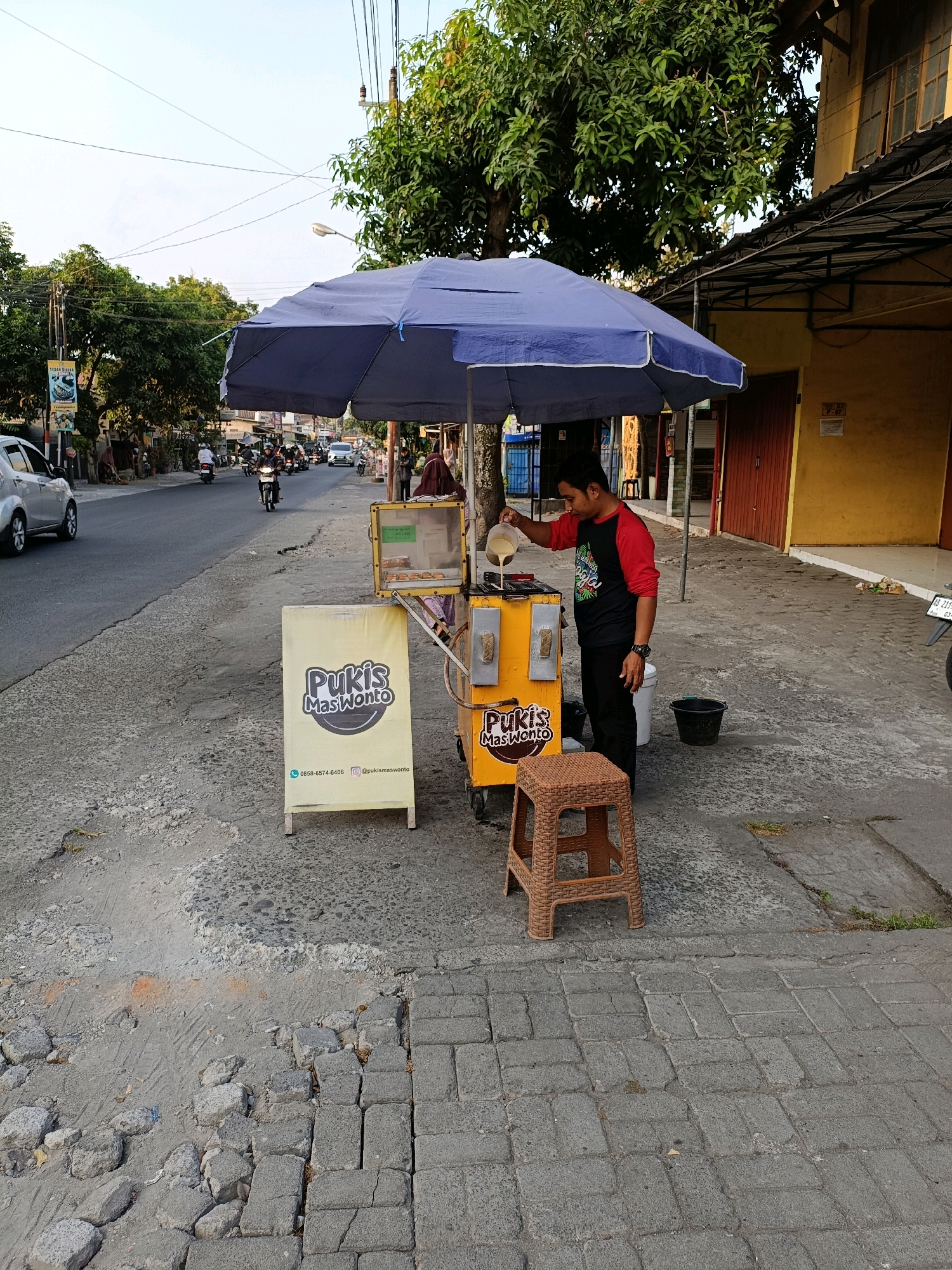 A man is pouring batter from a jug into a Pukis Mas Wonto food cart. The cart is yellow with a blue umbrella overhead. A sign with the Pukis Mas Wonto logo is propped in front of the cart. A brown plastic stool is next to the cart. The scene takes place on a street in Purwomartani, Indonesia, with a road, sidewalk, trees, and buildings visible. Several motorcycles and a car are on the road in the background.