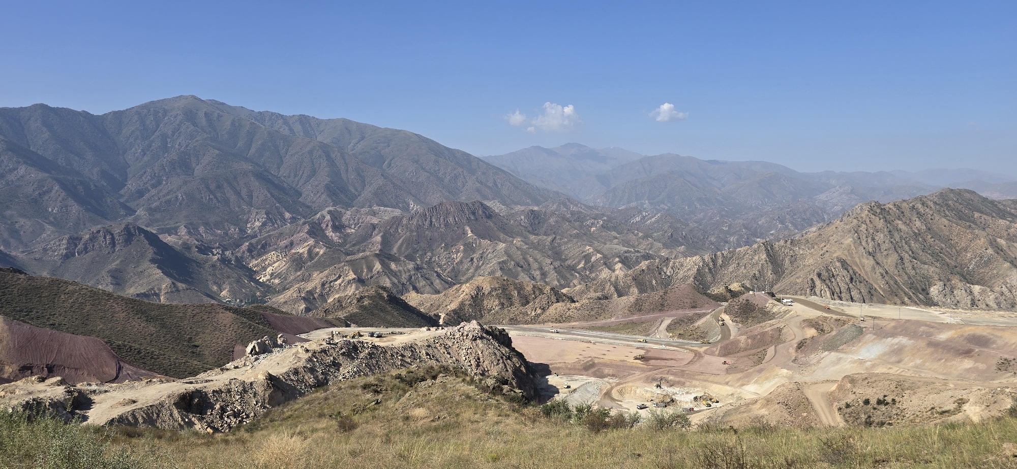 The image shows a vast mountain landscape under a clear blue sky. In the foreground, dry grass covers the rolling hills. The midground reveals a large-scale mining operation with extensive excavation and terracing on the mountainside. Several heavy machinery vehicles, including excavators and trucks, are visible working on the site. People, appearing as small figures, are also present on the excavation platforms. The excavated areas display layers of reddish-brown and gray soil. In the background, numerous mountain ranges stretch towards the horizon, with some peaks shrouded in haze. The overall scene depicts a combination of natural mountainous terrain and industrial activity. The location is Ak-Terek, Kyrgyzstan.
