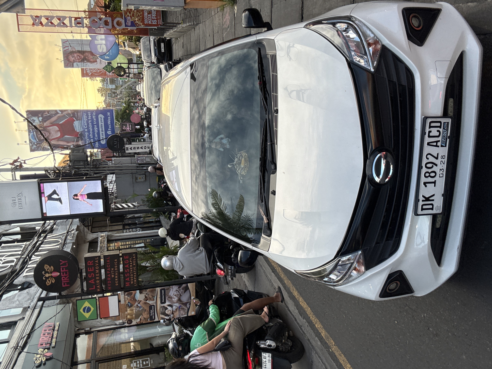 The image shows a street scene in North Kuta, Indonesia, with a white car prominently displayed in the foreground. The car's license plate reads "DK 1892 ACD" with a March 2028 expiration. Reflected in the car's windshield is the surrounding environment, including palm trees and buildings.
Along the street, there are several motorcycles parked, some with people on them. One person is wearing a green shirt and another a gray hoodie. Several businesses line the street, including "FIREFLY," which offers laser hair removal, hydrafacial with dermalogica, and massage. A Brazilian flag is visible on the storefront.
In the background, there are several billboards advertising various services, including pizza and a Canggu traffic guide. Overhead power lines and streetlights are also visible. The sky is visible in the background with a sunset. The overall scene suggests a busy commercial area in North Kuta.