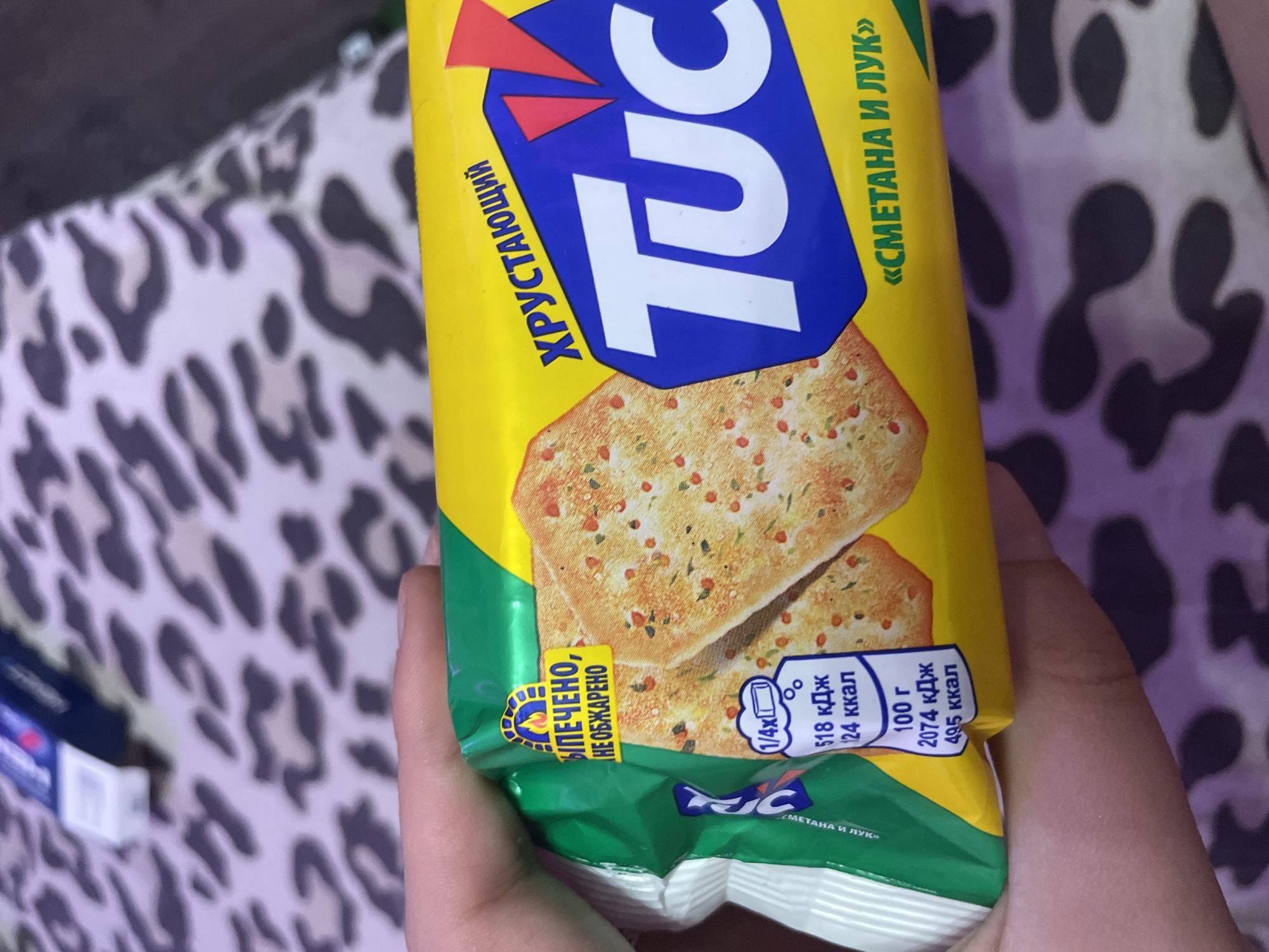 The image shows a close-up of a hand holding a package of TUC crackers. The crackers appear to be flavored with "sour cream and onion" in Russian. The packaging is yellow with blue and red accents. There is text on the packaging that says "ХРУСТАЮЩИЙ TUC" which translates to "CRUNCHY TUC". There is also nutritional information displayed in a small white box, showing that 100 grams of the crackers contain 495 calories and 2074 kJ. The background of the image is a blurry leopard-print fabric. The specific location in Vaganovo, Russia, cannot be determined from this image.