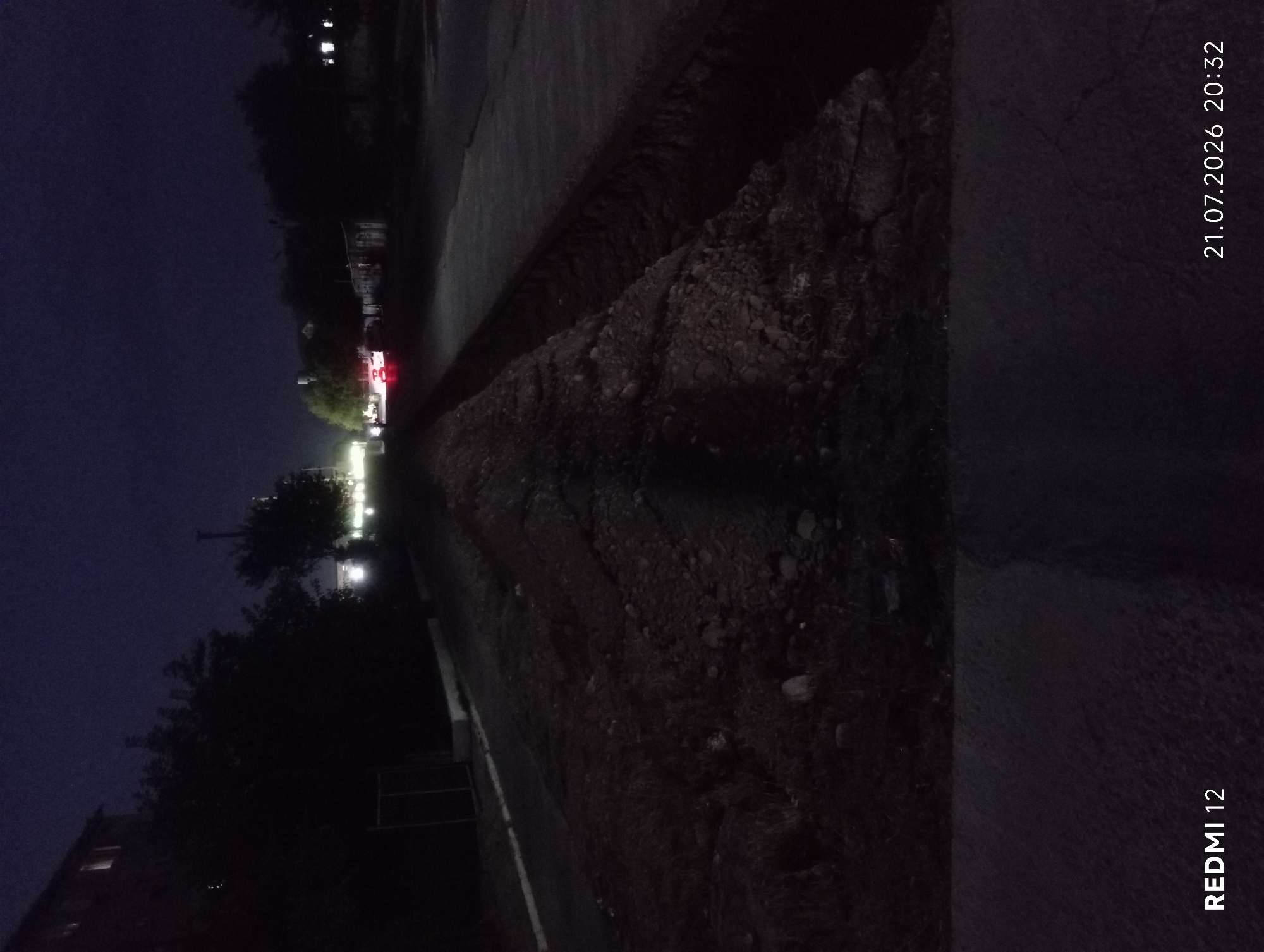 The image depicts an outdoor scene at night, likely in a residential or suburban area of Bekabad, Uzbekistan. A paved pathway runs diagonally across the frame, bordered on one side by a dark, uneven embankment covered in loose soil and rocks. On the other side of the pathway, there is a dark, blurry area with some faint lights visible in the distance, suggesting buildings or streetlights. Trees are silhouetted against the dark sky, their foliage rendered in shadow.
In the far distance, a brightly lit structure, possibly a shop or a building with illuminated signs, stands out with red and white lights. There are also some streetlights visible, casting a soft glow on the surroundings. The overall atmosphere is quiet and still, typical of a nighttime setting.
The image is oriented vertically and contains two instances of visible text. In the upper right corner, oriented vertically, is "21.07.2026 20:32", indicating the date and time the photo was taken. In the lower right corner, also oriented vertically, is "REDMI 12", which is likely the model of the phone used to capture the image. The weather appears to be clear, with no visible signs of rain or fog.