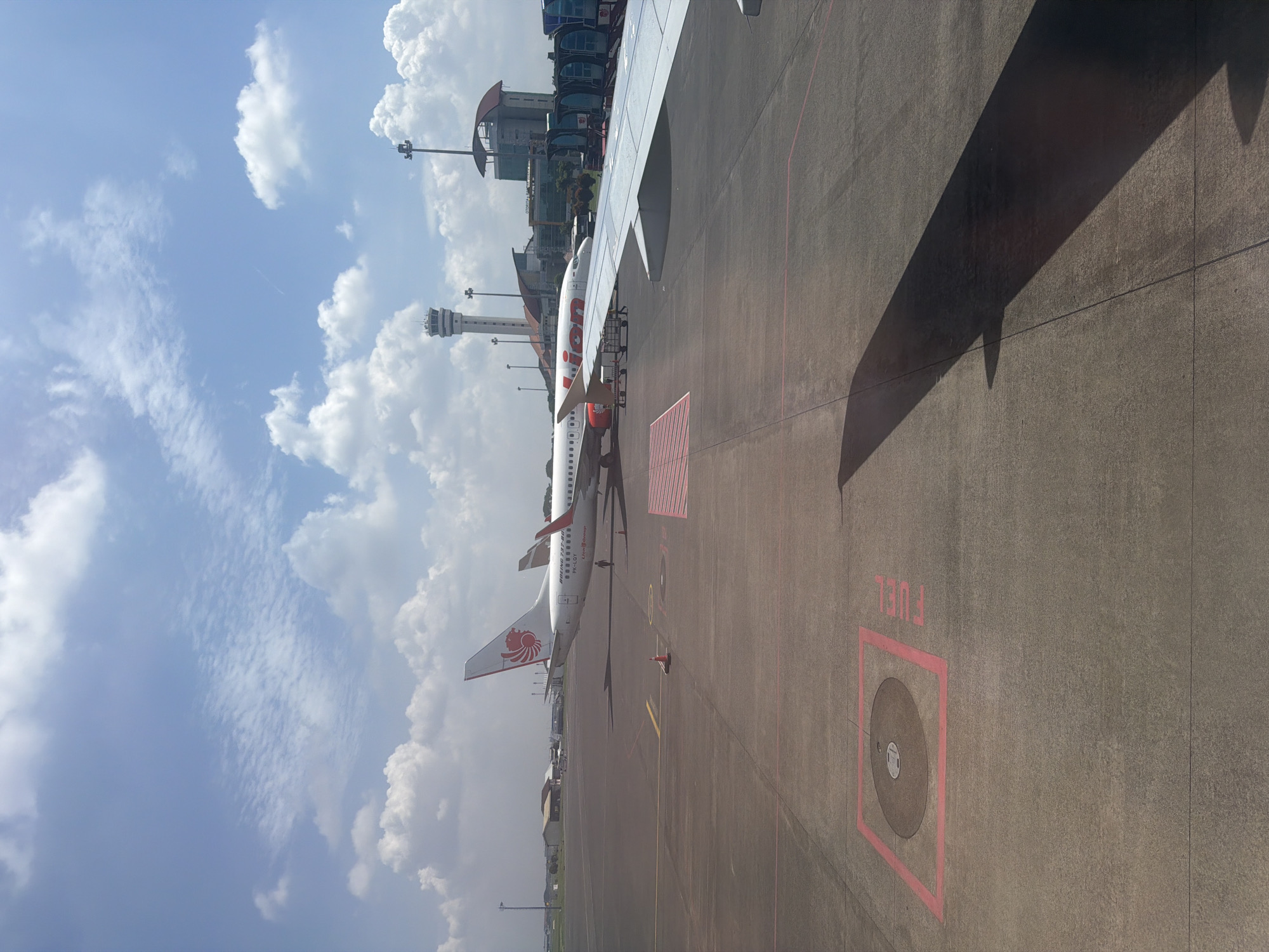 The image depicts a bright daytime scene at an airport, likely viewed from a terminal window or another aircraft. The primary subject is a white Lion Air Boeing 737-800 aircraft, bearing the registration PK-CLQ, parked on the asphalt tarmac. Its tail displays the distinct red and white Lion Air logo (a lion's head), and "Lion Air" is prominently written on its fuselage.
In the background, a tall, white air traffic control tower stands against a clear blue sky dotted with scattered, fluffy white cumulus clouds. Modern airport terminal buildings with reflective glass facades are also visible behind the parked aircraft.
The foreground shows the grey tarmac with various ground markings, including red and white stripes. A notable detail is a red rectangular outline on the tarmac, enclosing a circular metal cover, with the word "FUEL" painted nearby in red. The shadow of an aircraft wing, presumably the one the viewer is on or looking from, stretches across the right side of the tarmac, showcasing distinct shadow shapes of engine cowlings.
The overall setting is an active airport apron, indicating fair weather conditions. No specific individuals are clearly visible, but the scene suggests the regular operations of an airport, with the Lion Air plane potentially undergoing ground services or awaiting departure/arrival in Segoro Tambak, Indonesia.