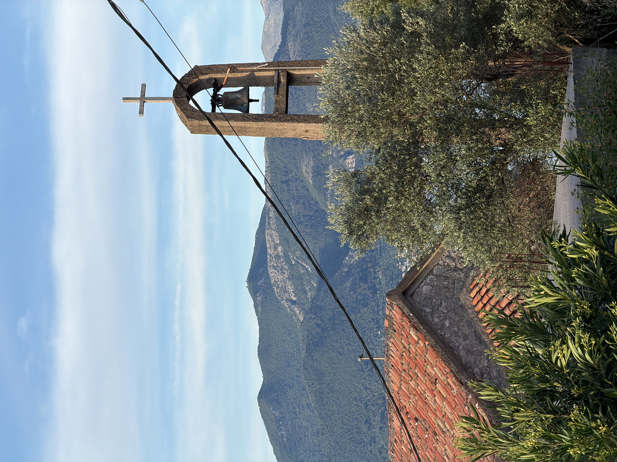 The image depicts a bell tower with a cross on top, situated in the city of Krašići, Montenegro. The tower is made of stone and houses a bell within its structure. In the background, a mountainous landscape is visible under a partly cloudy sky. An olive tree and other foliage partially obscure the view of a red-tiled roof of a building near the bell tower. Electrical wires run across the foreground of the image, slightly obstructing the view of the bell tower and the landscape beyond.