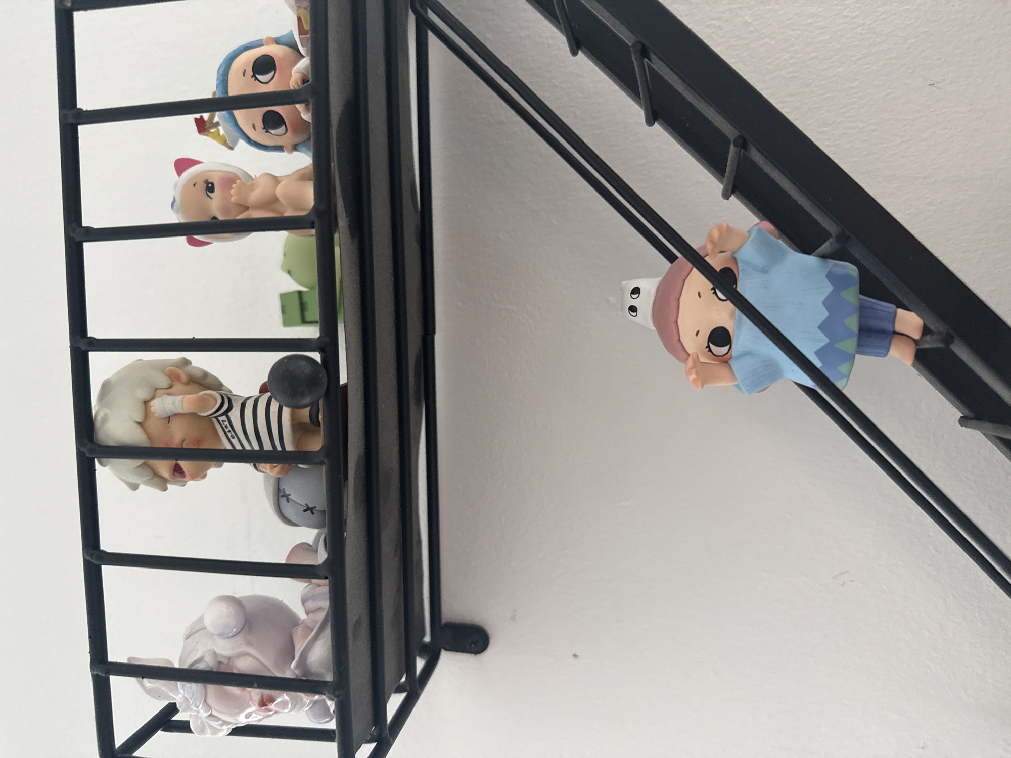 A low-angle close-up shot shows a black metal shelf affixed to a white wall. Inside the shelf, various small figurine dolls are visible. One doll, with a blue outfit and brown hair, appears to be hanging off the side of the shelf.