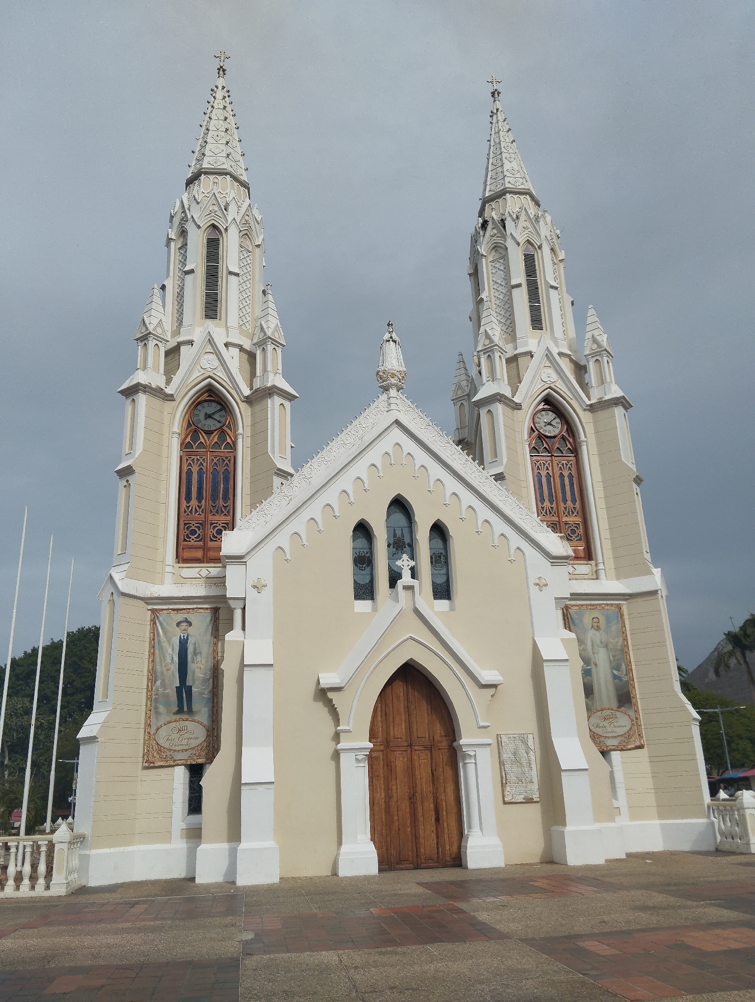 The image depicts the front facade of the Basilica of Our Lady of El Valle, a Catholic church located in El Valle del Espiritu Santo, Margarita Island, Venezuela. The church features a beige and white color scheme, with two tall, ornate towers flanking a central entrance. Each tower has a large, arched window with a clock above, and is topped with a spire. The central facade includes a large wooden door, framed by an archway, and several arched windows above. Banners with portraits hang on either side of the entrance. A low white railing runs along the front of the church, and the foreground shows a paved area. The sky is overcast. Green vegetation is visible in the background on either side of the church.