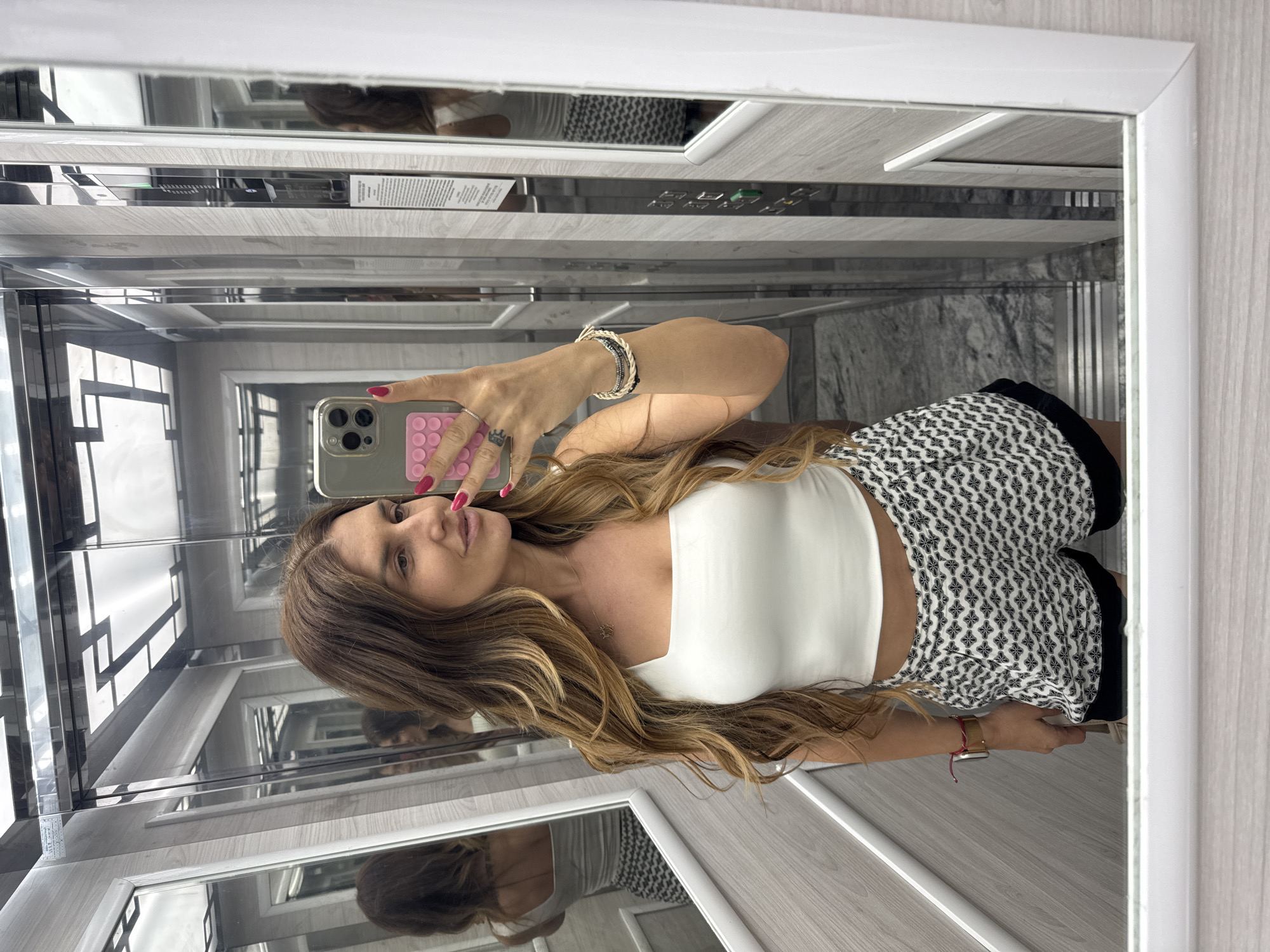 In an elevator with mirrored walls and a faux wood paneling, a woman with long, wavy brown hair and fair skin takes a selfie. She is wearing a white crop top and black and white patterned shorts. Her left hand, adorned with several bracelets and rings, holds a smartphone with a pink pop socket. Her right hand is extended, her nails painted red, as she holds the phone to capture her reflection. The elevator's interior features a control panel with illuminated buttons and a display screen on the right wall. Reflections of the woman and the elevator's interior are visible in the surrounding mirrors. The floor appears to be a grey marble pattern.