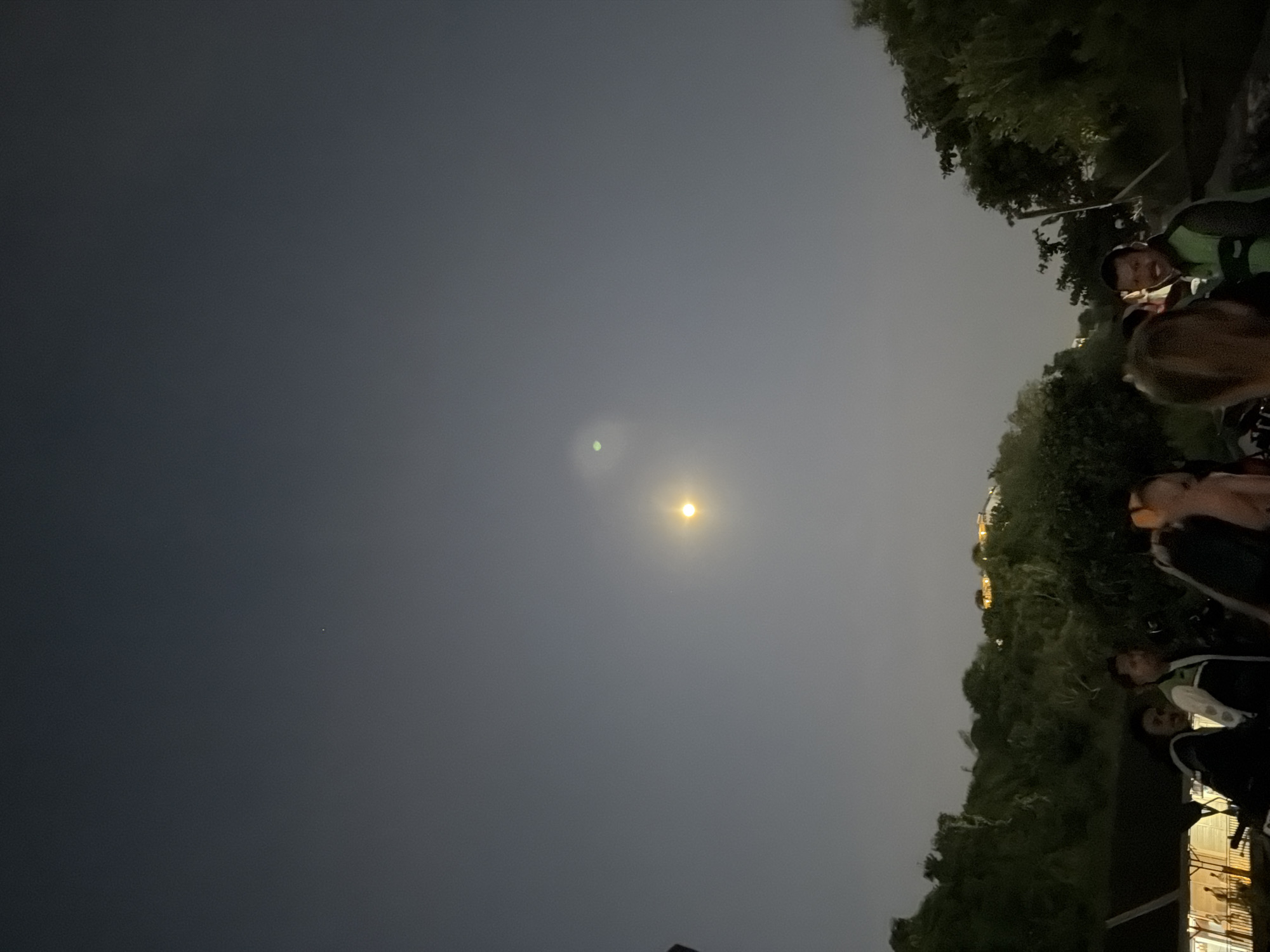At night in Kuta Selatan, Indonesia, the sky displays a gradient from deep gray to lighter gray near the horizon. Two celestial objects are visible: a faint green spot and a brighter, spherical light source. Silhouetted trees and vegetation line the top and right edges of the frame. A group of people is gathered near the bottom right, with some partially visible and engaged in conversation. Some artificial lights from buildings in the background are visible in the bottom right. The scene is oriented sideways with the sky dominating the upper left.