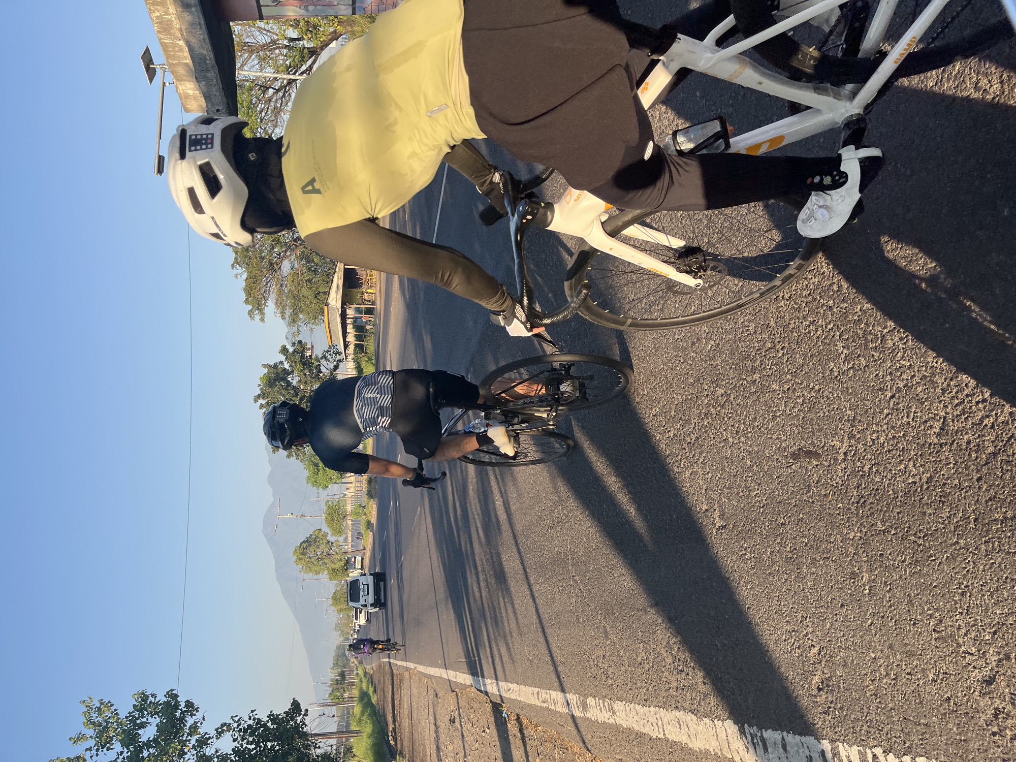 The image depicts two individuals cycling on a paved road, likely in Siring, Indonesia, under a clear blue sky during the day. The perspective is from behind the cyclists, showing their backs as they move forward. The cyclist in the foreground is wearing a white helmet, a yellow long-sleeved cycling jersey with the letter "A" prominently displayed, and black cycling shorts. They are on a white bicycle. The cyclist in the background is wearing a dark helmet and a dark and white striped short-sleeved cycling jersey with black shorts, riding a dark-colored bicycle.
The road is asphalt, with a white line marking the lane divider. To the left of the road, there is a grassy shoulder with some trees and utility poles. In the distance, on the right side of the road, a small structure with a yellow roof is visible, along with some trees and a concrete structure with a solar panel on top. Further in the background, a mountain range is visible under the bright sky. Several vehicles, including a white SUV and other cars, are on the road ahead of the cyclists. The sun is casting long shadows from the cyclists and trees across the road. The overall scene suggests an outdoor recreational activity during daylight.