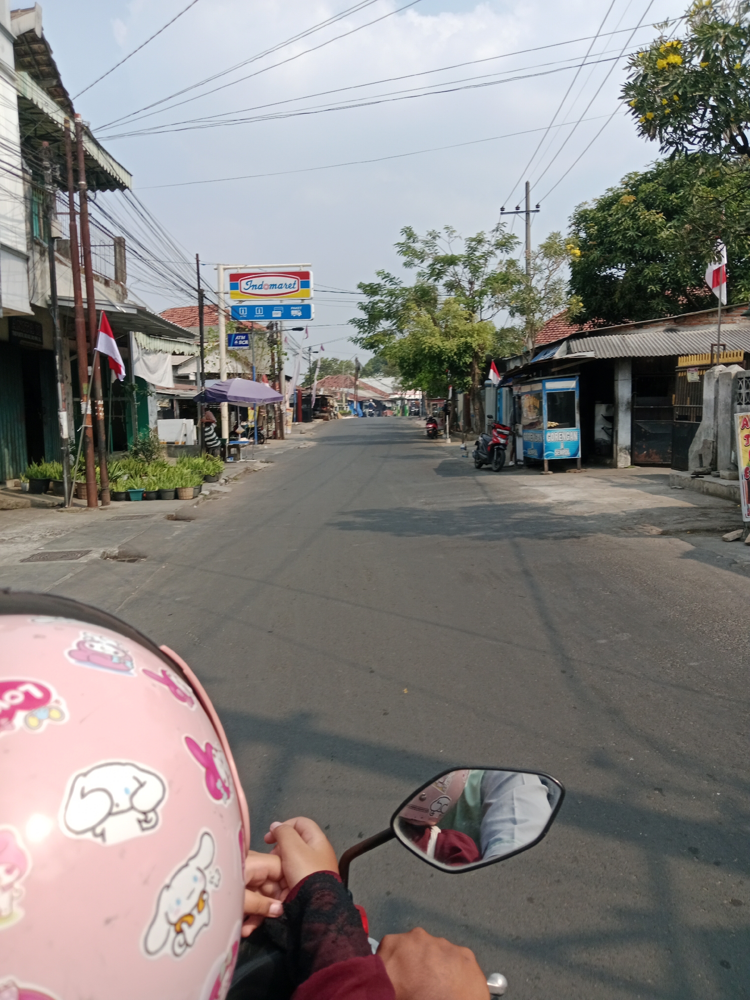 An eye-level, full shot captures a street scene in Kediri City, Indonesia, as viewed from a motorcycle. The road, paved and showing signs of wear, stretches forward into the distance, lined on both sides by low-rise buildings and shops. Overhead, a network of electrical wires crisscrosses the sky, and a bright blue sky with scattered clouds forms the backdrop. On the left, a building with white siding and dark window frames features a covered awning. Red and white Indonesian flags are visible, fluttering from poles. Potted plants are arranged in front of some of the buildings. A prominent Indomaret sign, a popular convenience store chain in Indonesia, is mounted on a pole above the street, displaying its distinctive red and blue logo. Further down the street, a food stall with a blue exterior and the word "GORENGAN" (fried food) is set up, with a motorcycle parked nearby. On the right, more buildings with corrugated metal roofs are visible, along with another Indonesian flag. The foreground is dominated by a pink helmet adorned with cartoon characters, which belongs to the rider. A hand, wearing a maroon sleeve with black lace trim, is visible on the handlebars. The mirror of the motorcycle reflects the rider, who is wearing the pink helmet, and a passenger or the back of the rider's head. The scene depicts a typical day on a local street in Indonesia, characterized by the presence of local businesses, residential structures, and everyday transportation.
