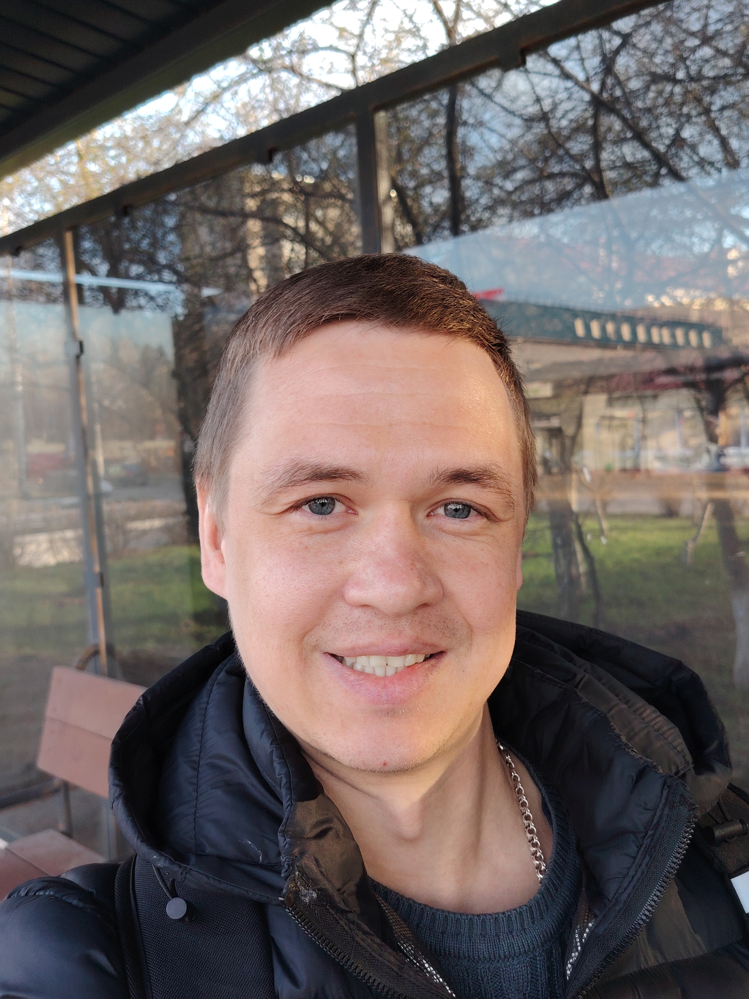 A smiling man, likely in his 30s or 40s, is captured in a close-up selfie, looking directly at the camera. He has short brown hair, blue eyes, and a fair complexion with light stubble. He is wearing a black puffer jacket with a hood, and a dark grey or black knitted sweater underneath, from which a silver chain necklace is visible.
The scene appears to be at a bus stop or shelter with prominent glass walls. The background is dominated by reflections on this glass, showing a bright, possibly overcast, sky and the intricate, bare branches of trees. Through the reflections and the glass itself, distant urban or semi-urban elements can be glimpsed, including indistinct buildings and patches of green grass. A wooden bench is visible in the lower-left background, suggesting an outdoor public space. The lighting is bright and natural, indicating daytime, possibly late autumn or early spring given the bare trees. The man's expression is friendly and cheerful, with a wide smile revealing his teeth. There is no visible text in the image. This scene is set in Chaikovsky, Russia.