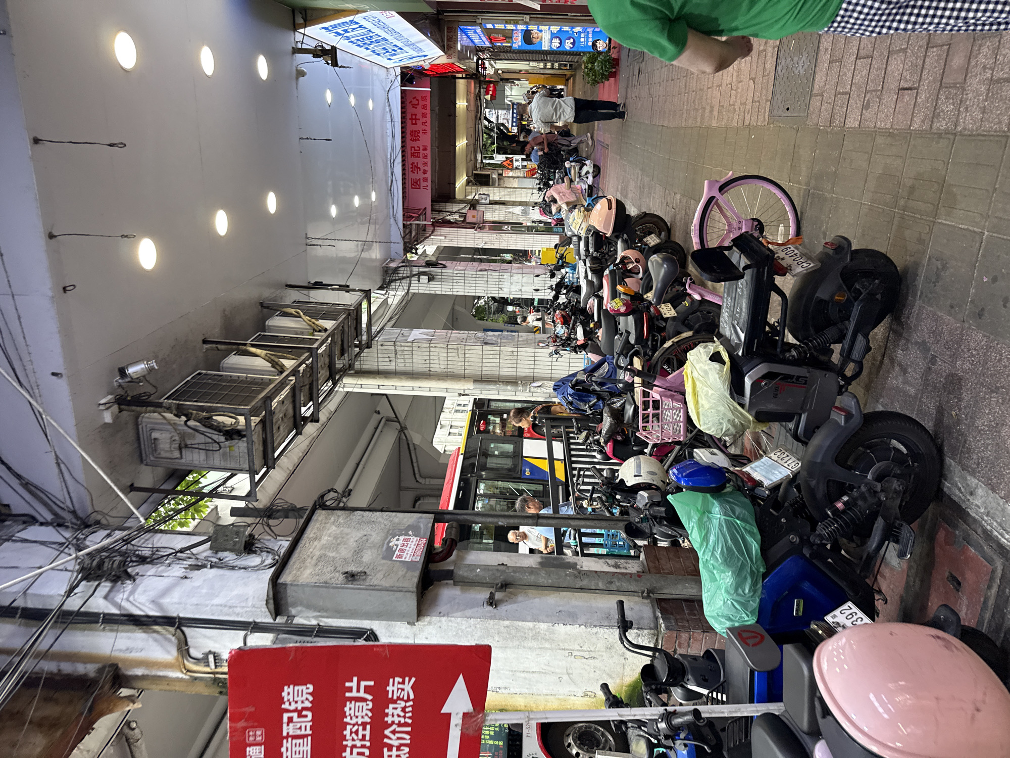This is a street scene in Yuexiu District, China, featuring a covered walkway lined with parked electric scooters and motorcycles. The walkway is illuminated by overhead fluorescent lights. On the left side, a red sign with white text, possibly an advertisement, is partially visible. Behind the sign, a bus is stopped at what appears to be a bus stop. Several individuals are present, some standing near the bus and others walking along the walkway. The ground is paved with brick-like tiles. Overhead, electrical wires are densely strung across the ceiling of the walkway. The atmosphere is urban, with a mix of commercial activity and transportation infrastructure.
