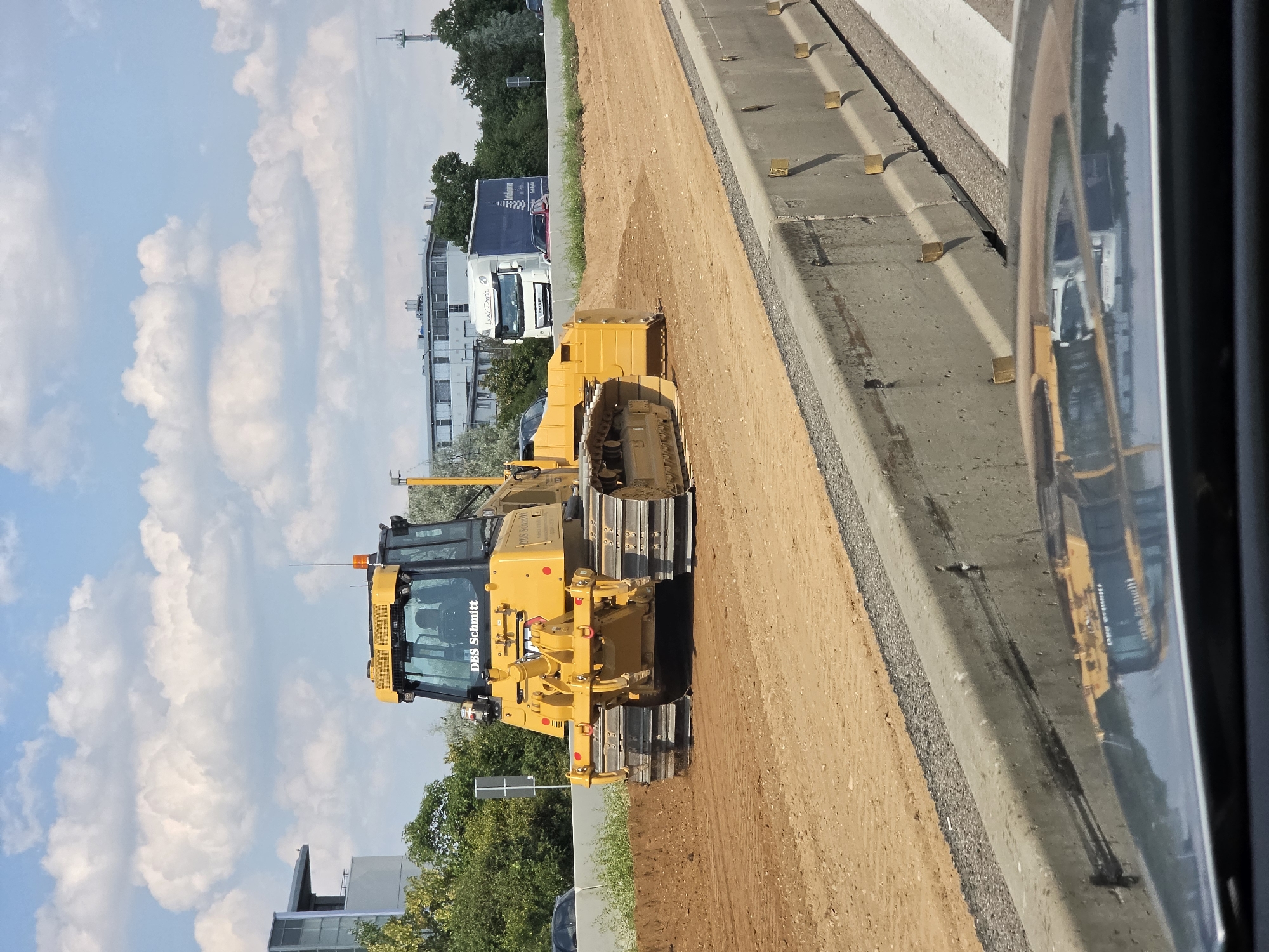 The image shows a yellow bulldozer with the text "DBS Schmitt" on its cab, operating on a dirt road adjacent to a concrete barrier. The bulldozer is positioned on the right side of the frame, with its tracks partially embedded in the sandy soil. The cab of the bulldozer is visible, featuring windows and what appears to be an orange warning light on top. In the background, there are trees and buildings, suggesting an urban or suburban setting. A white truck with a blue trailer is also visible further down the road, partially obscured by the bulldozer. The sky is bright blue with scattered white clouds, indicating it is daytime and likely sunny. The concrete barrier next to the bulldozer has yellow markers embedded in it at intervals. A reflection of the yellow bulldozer and surrounding scene is visible in a shiny surface to the right of the image, possibly a vehicle mirror. The image was taken in Rüsselsheim am Main, Germany. The scene appears to be a construction site, possibly related to roadwork or infrastructure development, given the presence of the bulldozer and the road surface. There are no visible people in the image. The overall impression is one of ongoing construction activity on a clear day.