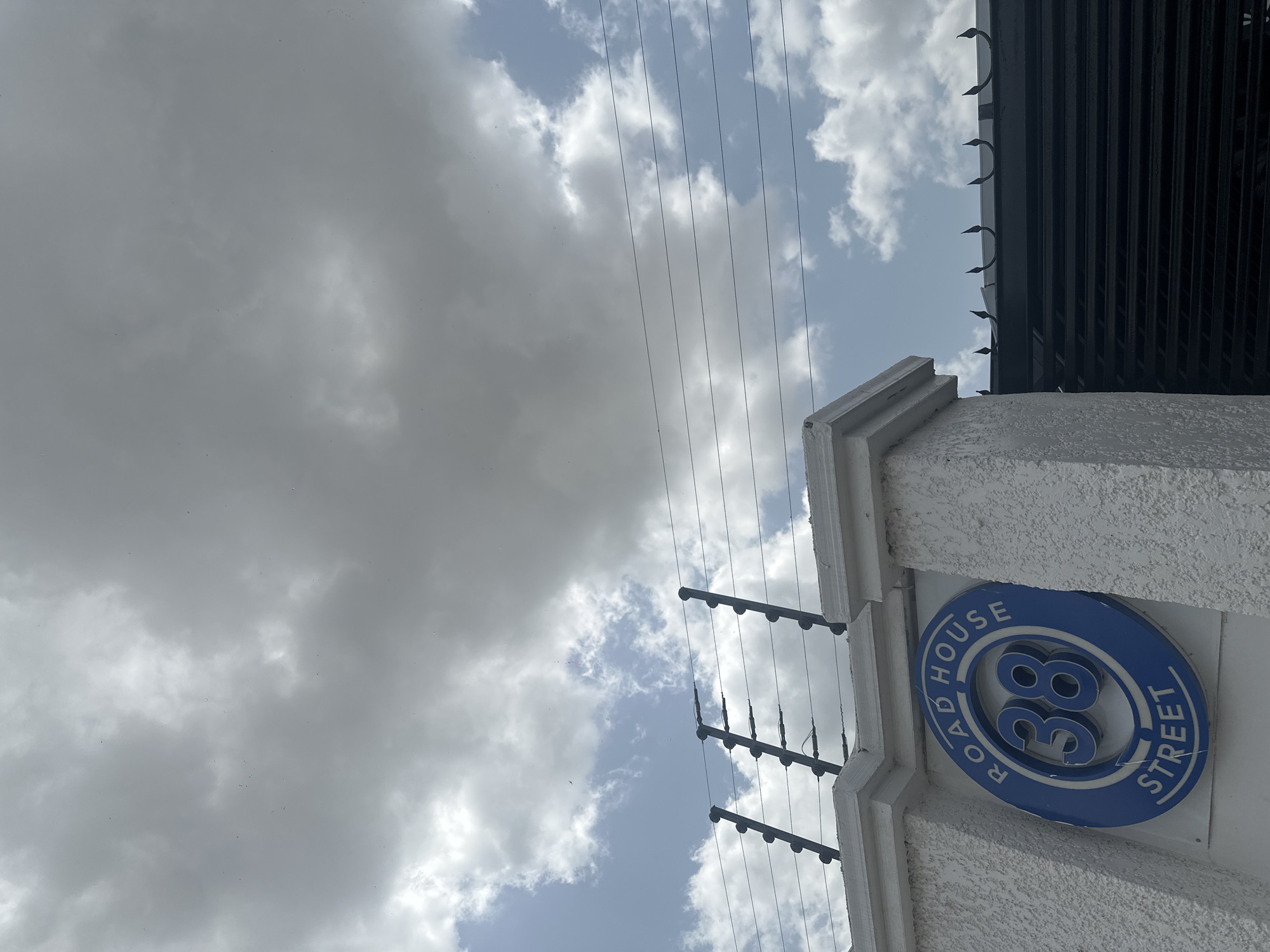 The image captures a view looking upwards, dominated by a dramatic sky filled with a mix of heavy, grey clouds and patches of bright blue sky. A security fence with multiple horizontal electrified wires is visible along the top of a textured white wall. Attached to this wall is a round blue sign with white lettering that reads "ROAD HOUSE 38 STREET". The sign is slightly tilted. To the right of the white wall, a dark, corrugated metal gate is visible, topped with decorative pointed spikes. The location is identified as Howayi, Nigeria.