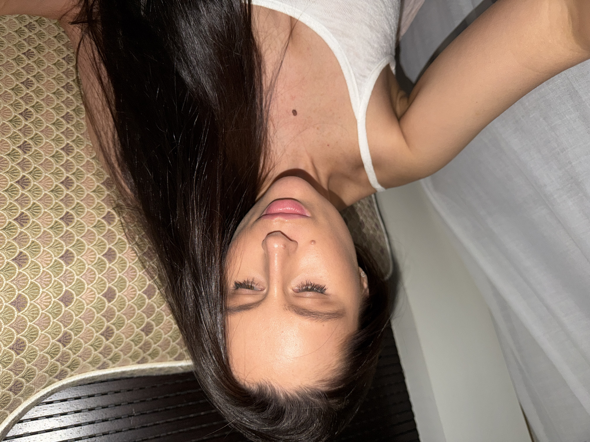A woman with long, dark hair is lying down, seemingly indoors. She is wearing a white tank top. A mole is visible on her upper chest/neck area. The background includes a patterned surface and a white curtain or sheet. The location in the city of Khimki, Russia, is not possible to confirm from the image.
