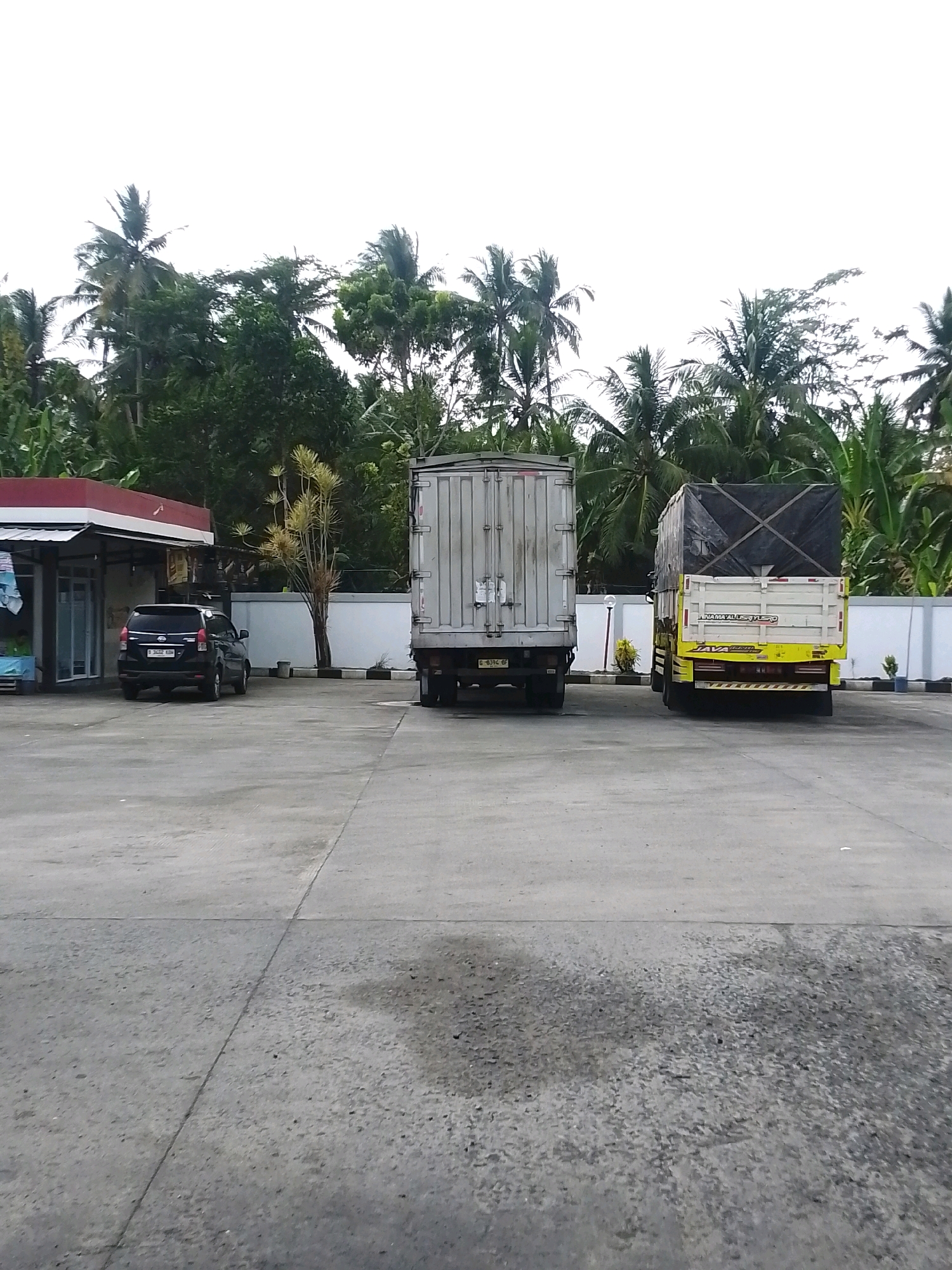 In Sukaresik, Indonesia, a black SUV is parked in front of a small building with a red awning. Two trucks are parked behind the SUV, a silver box truck to the left and a yellow and white truck with a black tarp to the right. The ground is paved with concrete, and in the background, there are numerous palm trees and other vegetation. The sky is overcast and white.