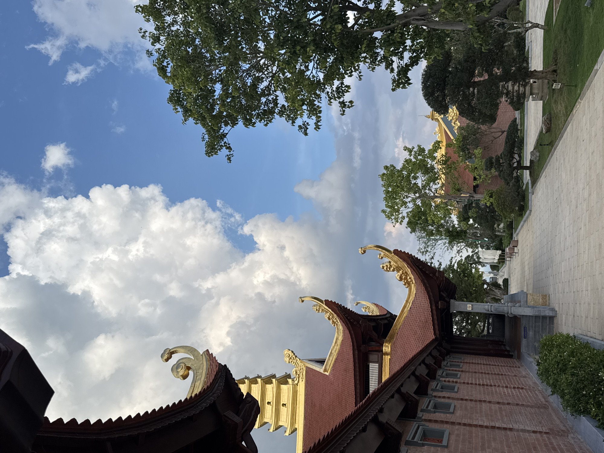 The image shows a view of a temple in Diên Khánh, Vietnam. The temple features ornate architecture with red tiled roofs and golden decorative elements on the eaves. A golden pagoda-like structure is visible in the background. The foreground shows a paved walkway lined with manicured trees and grass. The sky is a mix of blue with dramatic white clouds. The overall scene conveys a sense of tranquility and cultural richness.
