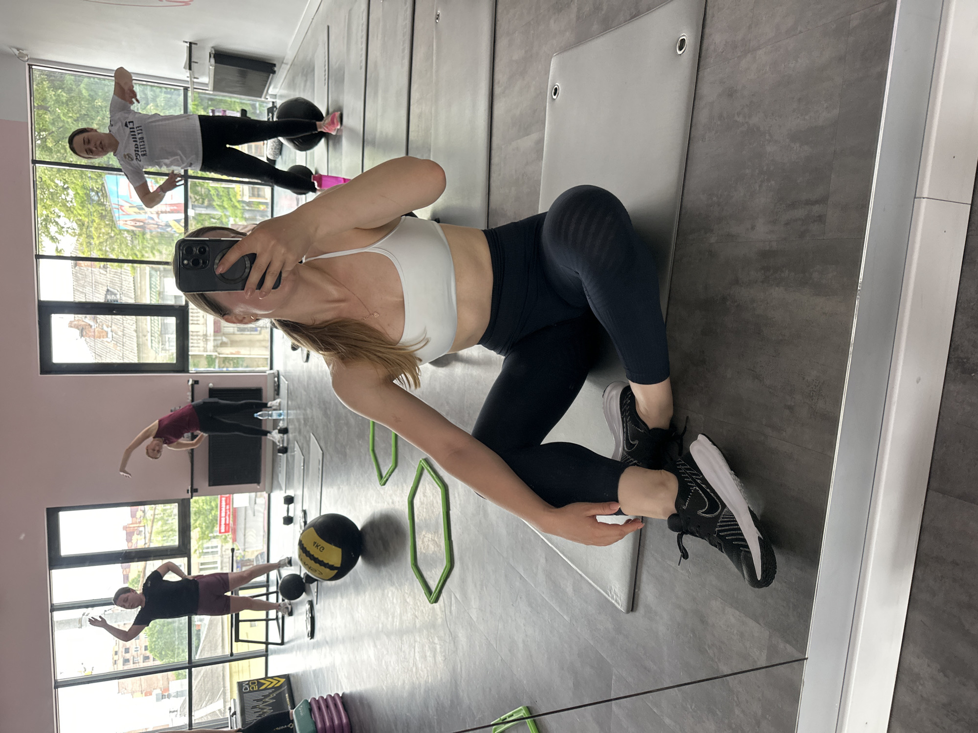 The image depicts a fitness studio located in Ternopil, Ukraine, captured as a mirror selfie. In the foreground, a woman is seated cross-legged on a grey exercise mat, wearing a white sports bra, dark blue leggings, and black Nike sneakers. She holds a dark-colored smartphone, partially obscuring her face, to take the photograph. A delicate necklace with a heart-shaped pendant is visible on her chest. Her dark hair is pulled back, with some strands falling over her left shoulder. The bottom edge of the large mirror features a white and silver frame.
Reflected in the mirror, in the midground, three other women are engaged in exercises. To the left, a woman in a black t-shirt and dark shorts performs an overhead arm stretch. Adjacent to her, a woman in a dark red t-shirt and black leggings leans to the side in a torso stretch. Further to the right, a third woman wearing a white t-shirt with mirrored text ("FLY EMIRATES" and "REAL MADRID") and black leggings stands with her arms slightly bent and extended outwards, wearing pink sneakers.
Exercise equipment is distributed across the dark grey tiled floor, including several grey exercise mats, a yellow and black medicine ball labeled "1KG," two small black dumbbells, and at least three green hexagonal agility rings. Two pink water bottles are also visible.
Through large windows along the studio's back wall, an urban outdoor scene is visible. This includes green trees, various buildings with residential architecture, and a partially visible colorful billboard or advertisement. The sky appears overcast. The interior walls of the studio are painted light pink at the top section and white below.