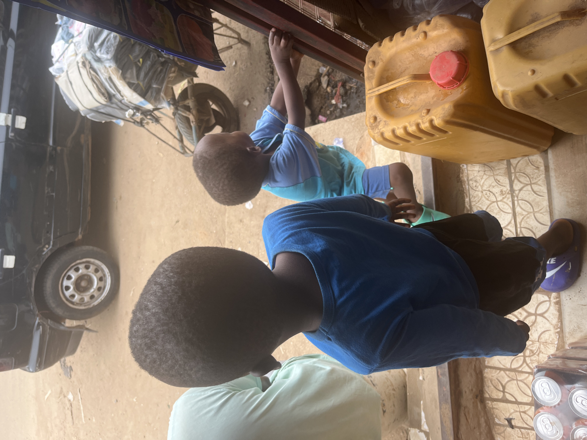 Two young boys are in the foreground of the image. The boy on the left, seen from behind, wears a blue long-sleeved shirt and dark shorts. The boy on the right, facing left, is wearing a light blue and darker blue short-sleeved shirt and blue shorts, with his hands on a red metal structure. He is partially obscured by the boy on the left.
In the background, a black vehicle is parked. To the right of the vehicle, a cart or wheelbarrow is loaded with what appears to be wrapped goods, possibly textiles or mattresses, secured with plastic wrap and straps. The environment suggests an outdoor setting, possibly a street or market area, with a dusty ground.
In the immediate foreground, to the right, are two large yellow plastic containers, one with a red cap. To the left and bottom, a pack of canned drinks is visible. The overall scene appears to be in a busy urban environment. The presence of the vehicle, cart, and market-like setup suggests a commercial or logistical area within the city of Abuja, Nigeria.