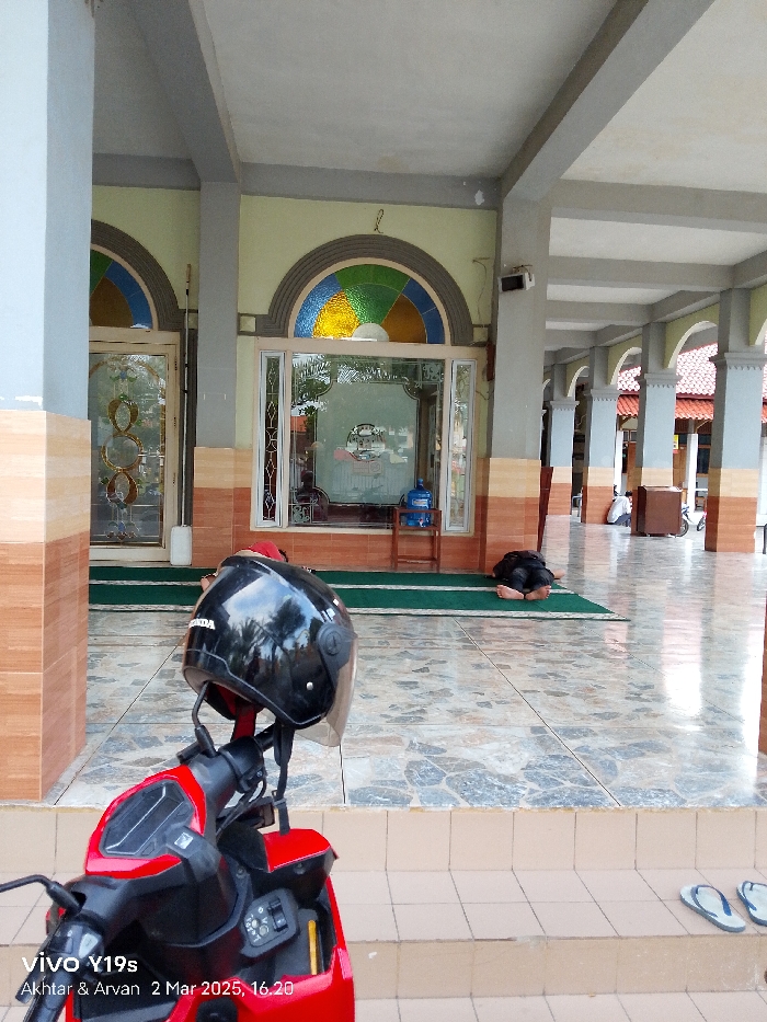 The image shows a red motorcycle parked in front of a building in Kecamatan Haurgeulis, Indonesia. The motorcycle is partially visible, with only the front wheel, handlebars, and a black helmet visible. The helmet is facing the camera and sitting on the handlebars. The building has a long, covered porch with gray tiled floors and multiple pillars. A man is lying on his stomach on the floor, with his head turned towards the right. The man is wearing a black shirt and pants. The porch is partially obscured by a large window, with a stained glass design, overlooking a garden area. The building is likely a mosque or religious building based on the architectural style and the presence of the man performing Islamic prayer on the floor. The image was taken at 16:20 on 2 March 2023.
