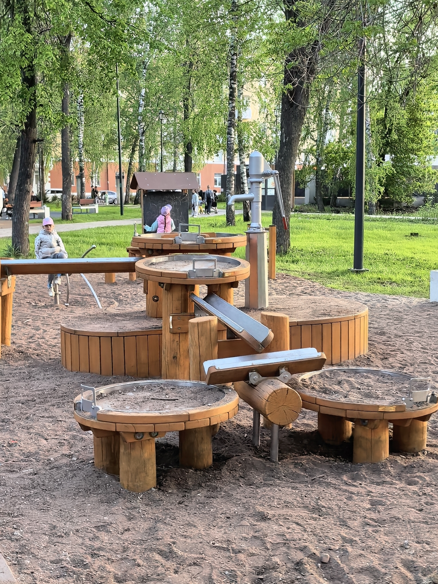 The image depicts a children's playground in Sterlitamak, Russia. The playground features various wooden play structures, including tables, platforms, and a water pump. Two young children are visible: one wearing a white jacket and pink hat is seated on a wooden structure, and another wearing a pink jacket and hat is standing on one of the platforms. The playground surface appears to be covered in sand. In the background, trees, grass, buildings, and other people are visible, suggesting a park or public space setting.