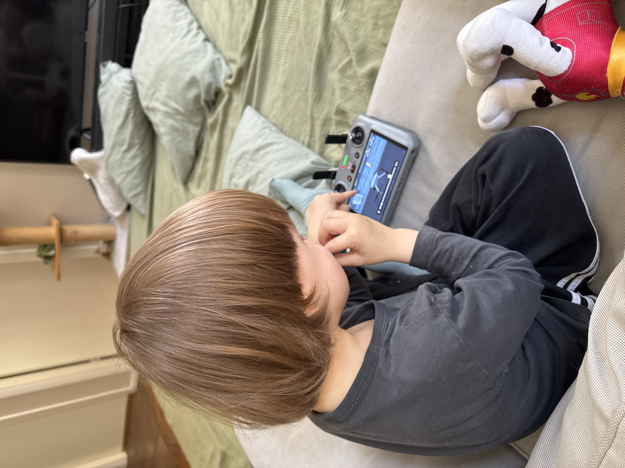 A child with short brown hair is sitting on a beige sofa, looking at a gray drone controller and touching its screen. The child is wearing a gray long-sleeved shirt and black pants with white stripes on the side. Next to the child is a stuffed animal depicting a dog wearing a red vest. Behind the child, there are light green pillows on the sofa. In the background, there is a TV, a wooden cat tree, and a wooden floor. The location cannot be determined from the image.