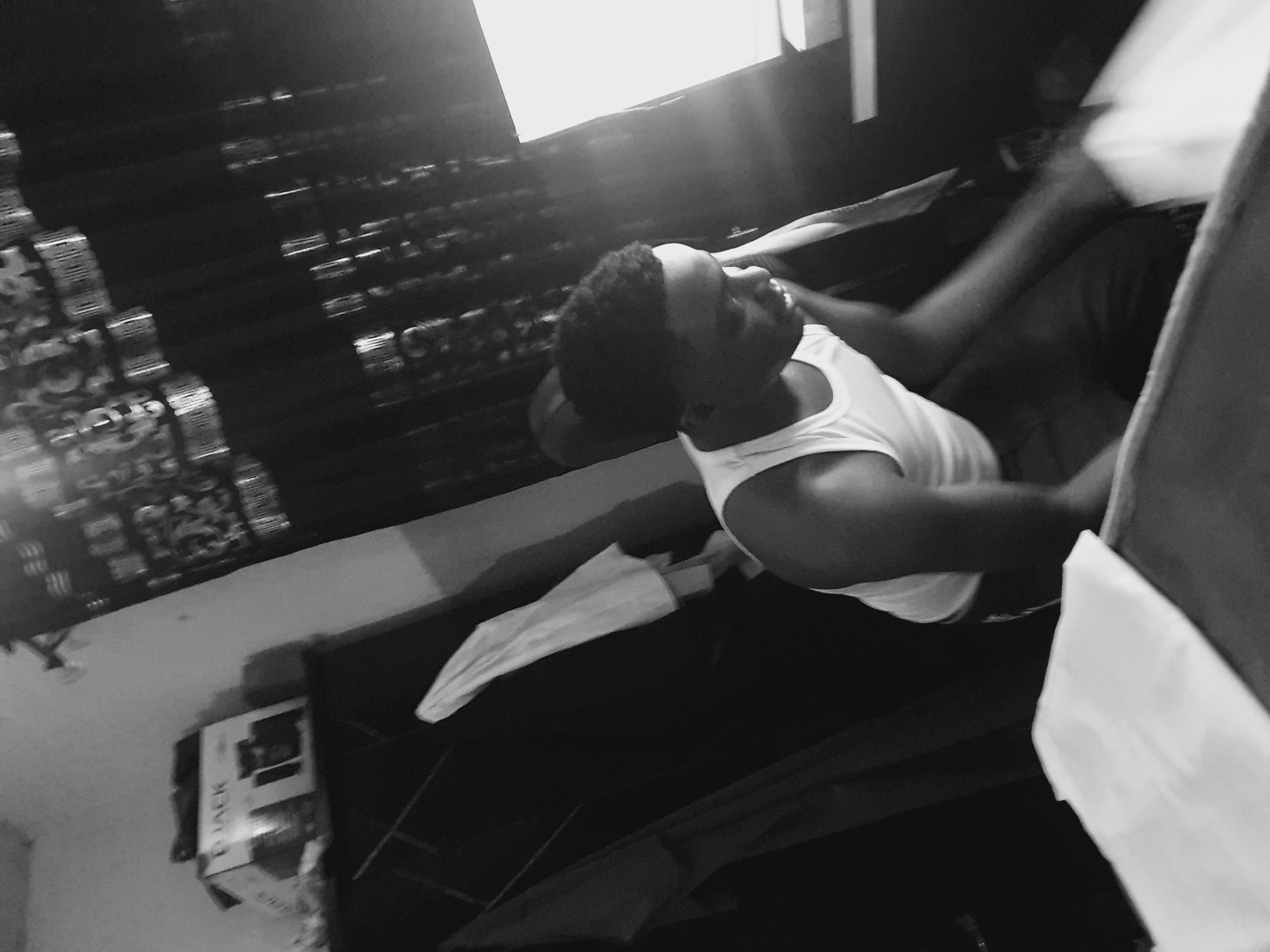 The image shows a person in a white tank top and dark shorts lying on what appears to be a bed. The background includes a window with blinds and a patterned wall. There is a box on the floor near the bed. The person appears to be smiling and looking upwards. The location, Lokoja, Nigeria, cannot be confirmed solely from visual evidence in the image.