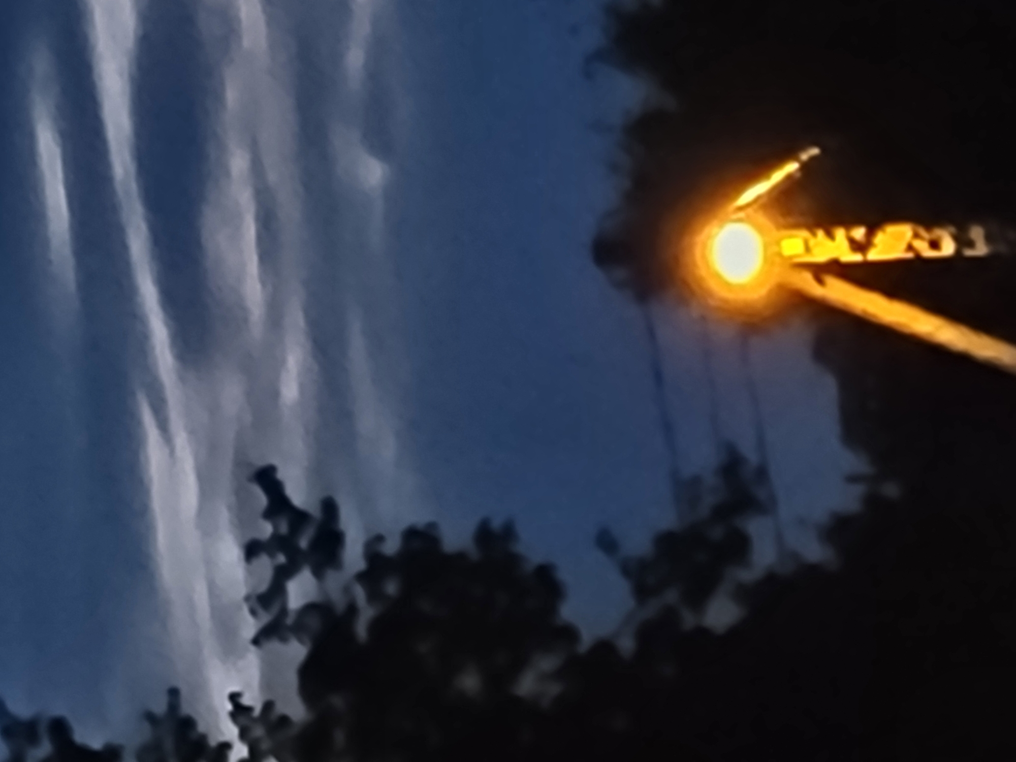 The image depicts a dark, atmospheric scene, likely taken at dusk or night, with a streetlamp in the foreground casting a warm, orange glow. The streetlamp appears to be a modern design, possibly a "park lamp" or "decorative lamp," judging by its structure and bright illumination. Behind the lamp, silhouetted trees are visible against a deep blue sky with streaks of lighter clouds, suggesting a twilight or overcast condition. The overall ambiance is somewhat moody and tranquil. The location is specified as Jelgava, Latvia, a city known for its historical architecture and parks, which could provide a context for such a scene. However, without specific landmarks or unique identifying features within the frame, pinpointing the exact location within Jelgava is not possible from the image alone.