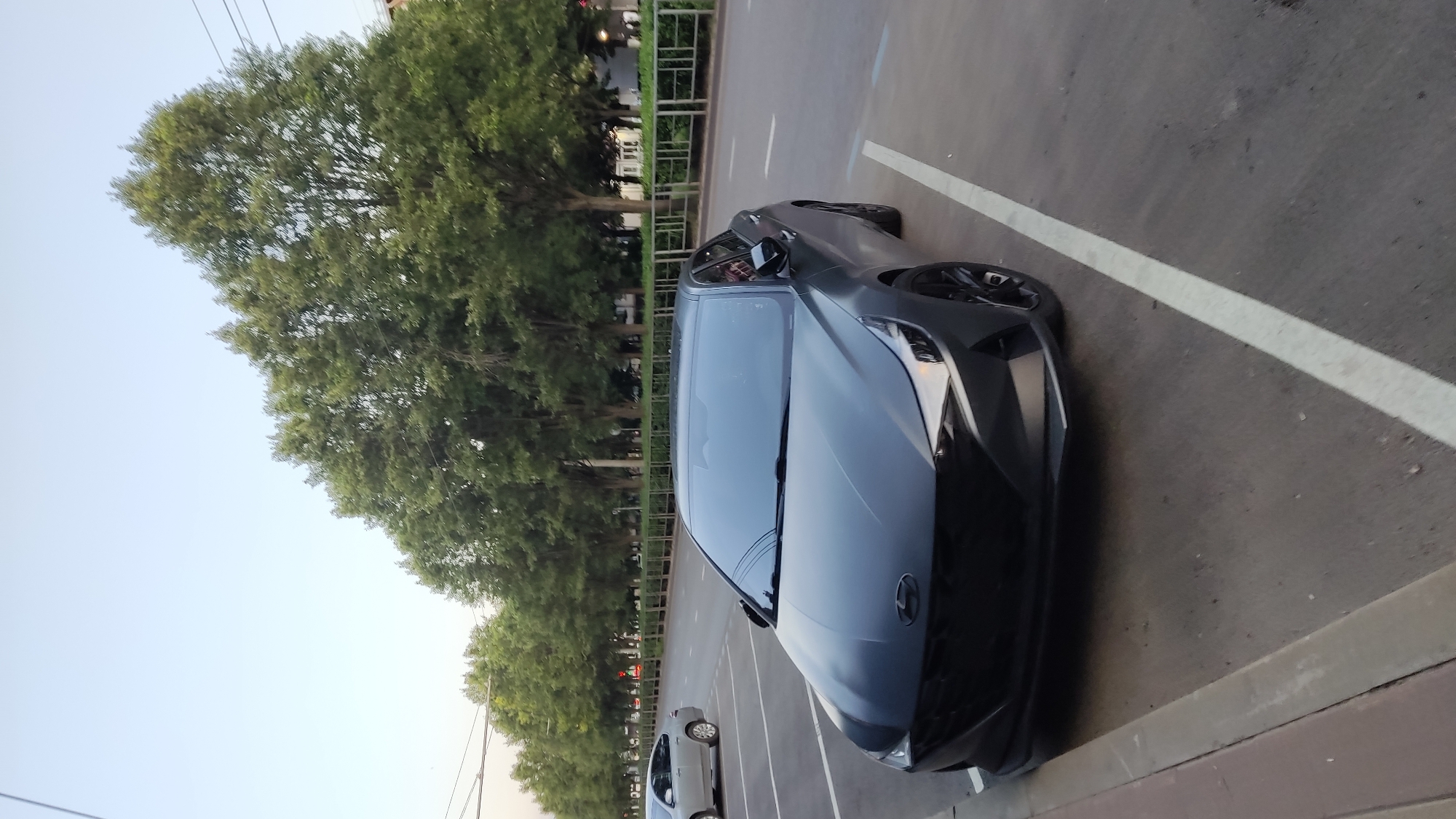 The image shows a dark gray Hyundai Elantra parked on a city street. The car is the primary subject, positioned in the foreground and angled towards the right. The street has a paved surface with white lane markings. To the left of the car, there is a curb and a sidewalk. In the background, a line of trees with dense green foliage stretches along the street, with a light-colored sky visible above. A light-colored sedan is parked further down the street on the left. The scene appears to be an urban environment, possibly in Krasnodar, Russia, given the mention of that location. The lighting suggests it might be late afternoon or early evening.