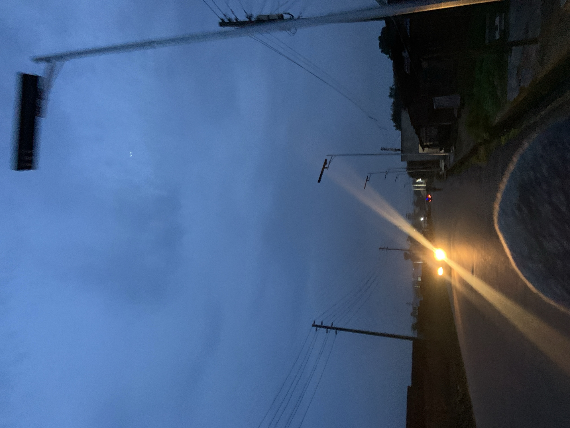 The image depicts a rural road scene at dusk or dawn in Barikin Jirga Summa, Nigeria. The sky is a deep, cloudy blue. A paved road runs through the scene, with a car's headlights illuminating the path and creating bright streaks of light. On the right side of the road, there are several buildings, appearing to be simple structures with dark roofing, and vegetation. Utility poles with multiple wires are visible along the road. A modern street lamp with a rectangular fixture is prominent on the right side of the image, and other street lamps are spaced along the road in the distance. The left side of the road features a dark, low-lying structure and more utility poles. A fuzzy, rounded object, possibly the top of a person's head or shoulder, is in the foreground, obscuring part of the road.