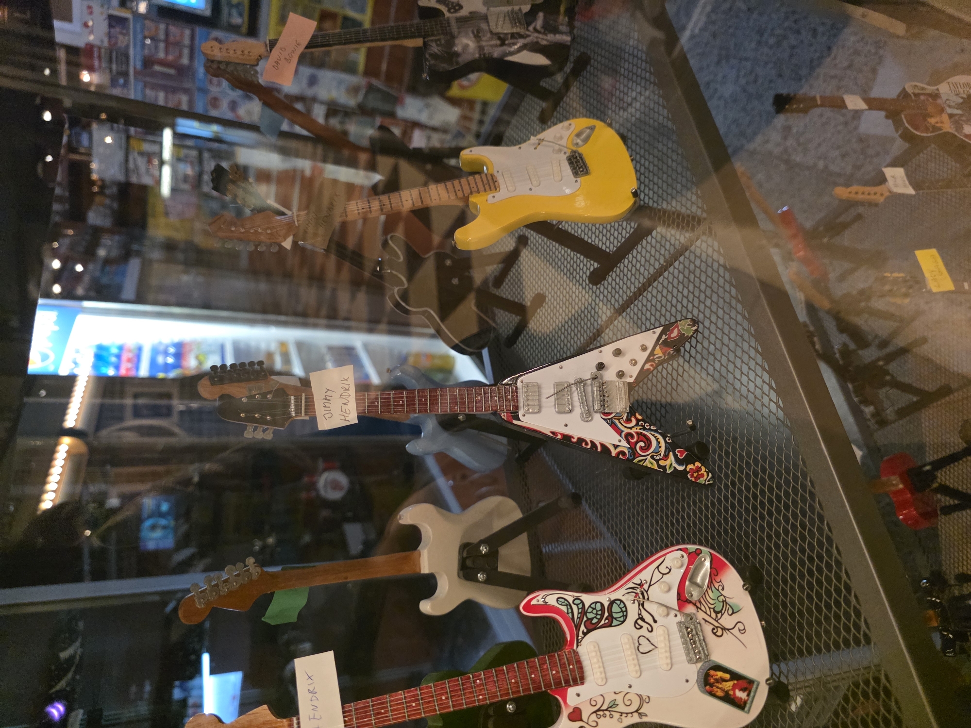 The image shows a display of miniature guitars in a store. The guitars are arranged on a tiered metal shelf. Several of the guitars are adorned with colorful designs, and some have labels attached, including "Jimi Hendrix" and "David Bowie." In the background, reflections show the interior of the store, which appears to be well-lit with various shelves and displays. The overall impression is of a retail environment showcasing musical memorabilia.