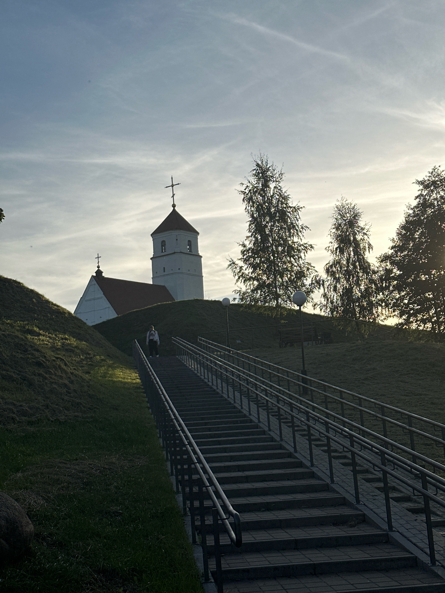 The image shows a person ascending a long outdoor staircase in the city of Zaslavl, Belarus. The staircase, made of stone pavers and bordered by metal railings, leads up a grassy incline. At the top of the incline, a white church with a red roof and a prominent bell tower is visible against a pale sky with wispy clouds. Several trees with sparse foliage are silhouetted against the sky on the right side of the frame. The scene appears to be illuminated by soft, natural light, possibly from a setting or rising sun, casting shadows on the grass and stairs.