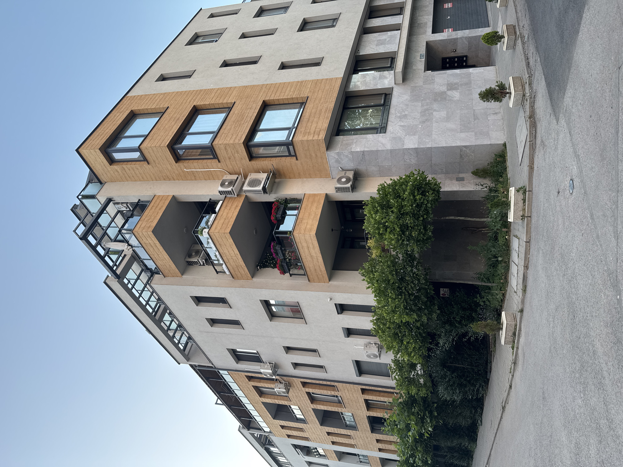 This is a street-level, eye-level, outdoor, daytime, full shot of a modern apartment building located in Sofia, Bulgaria. The building is multi-story with a contemporary architectural design, featuring a mix of light-colored stucco and wood-paneled facades. It has numerous windows, balconies with glass railings, and air conditioning units attached to the exterior. A glass-roofed structure is visible on the top level.
The building is situated on a street corner with a paved sidewalk running alongside it. Greenery, including trees and bushes, is planted along the sidewalk and in front of the building. Several small, potted evergreen trees are placed on the sidewalk near the building's entrance. The street in the foreground is paved with asphalt. The sky above is clear and blue. There are no people visible in the image. The scene appears to be a residential area in the city.