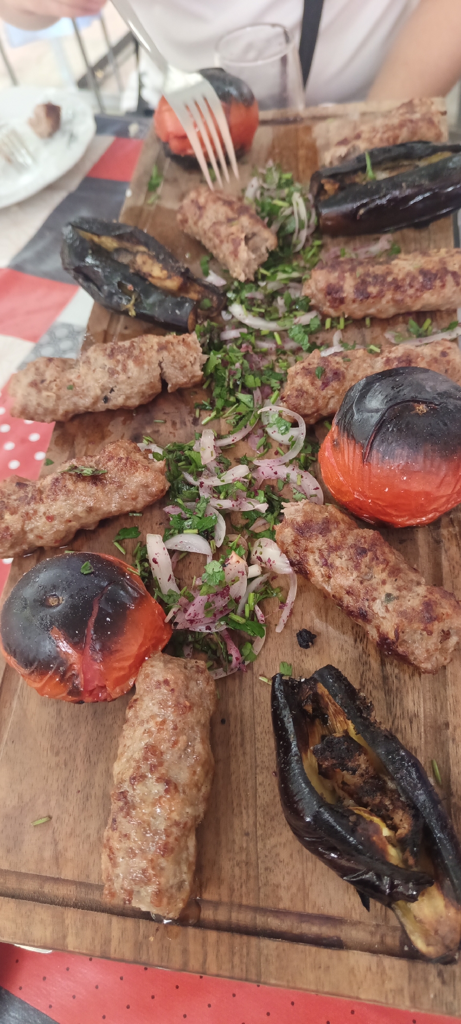 A top-down, medium shot shows a platter of grilled food, likely served at a restaurant in Khudat, Azerbaijan. The platter is made of wood and holds several pieces of grilled minced meat skewers (kebabs), whole grilled tomatoes, and halves of grilled eggplant. Chopped parsley and sliced red onions are scattered among the food as a garnish. A fork is visible in the upper right quadrant, piercing one of the grilled tomatoes, indicating that someone is in the process of eating. The background is blurred, but suggests a dining setting with a red and white checkered surface on the left and a red tablecloth with white polka dots on the bottom.