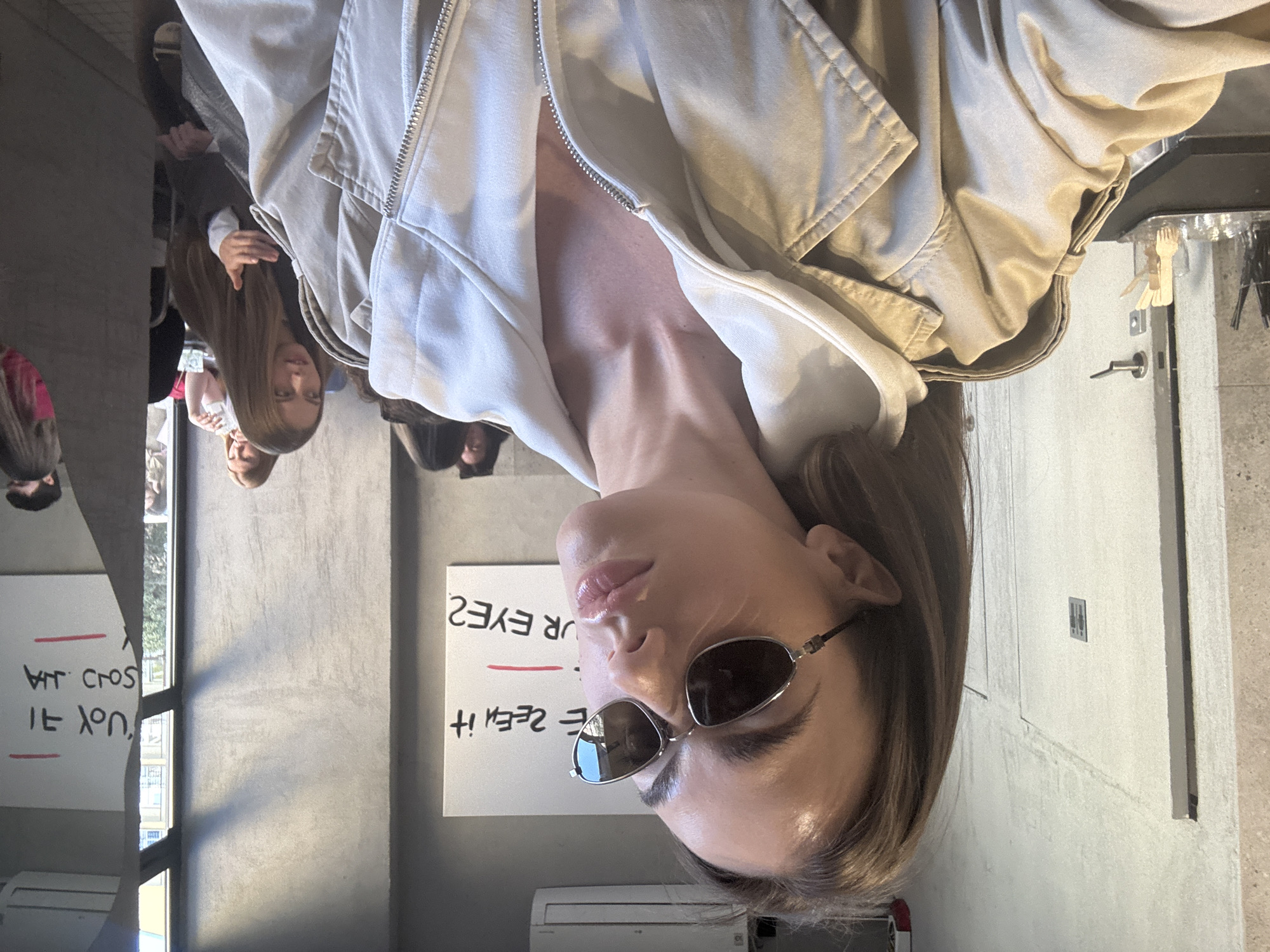 The correctly oriented image features a young woman in a medium close-up, situated in an indoor setting in Warsaw, Poland, during daytime. She is looking upwards and slightly to her left, her lips parted and glossy. She has medium-length brown hair and wears small, oval-shaped dark sunglasses. Her attire consists of a light-colored, possibly beige or off-white, zipped jacket, partially unzipped.
In the blurred background to her left, several other people, mainly young women with long hair, are visible, either congregating or reflected in a surface. To her right, a white board displays reversed handwritten English text: "YOUR EYES" underlined in red, and below it, "HAVE YOU SEEN IT". An air conditioning unit is located beneath the whiteboard. Further back on the right, a light-colored wall, a door handle, and part of a door are visible. A small, wooden hand-shaped decorative object hangs above the door. The overall scene suggests a casual atmosphere in a public or semi-public indoor space. The main subject's expression is neutral or contemplative.