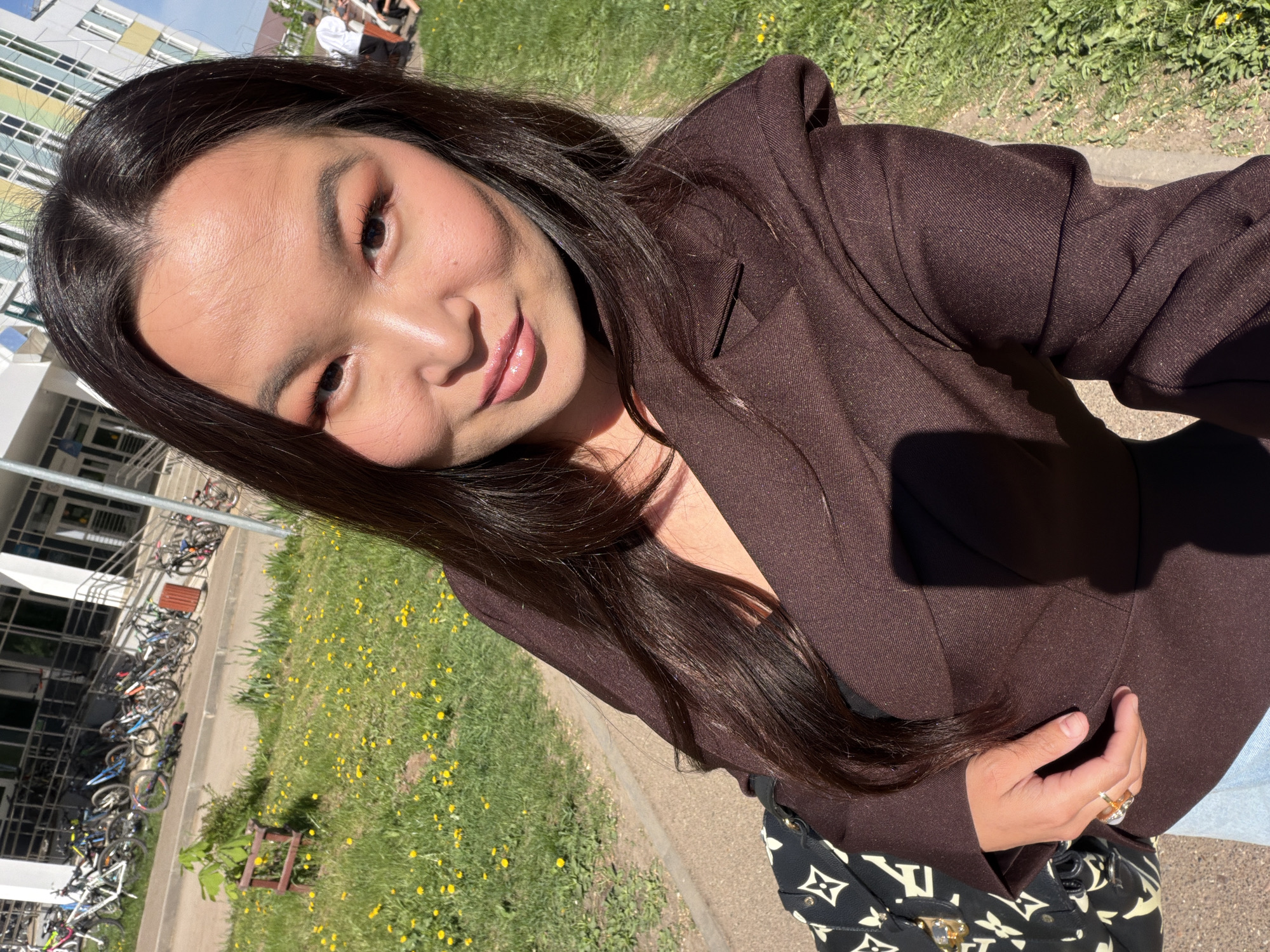 A medium-angle selfie shows a woman with long, dark hair, wearing a brown blazer. She has fair skin, dark eyes, and is smiling slightly. The background consists of a building, grass, and bicycles. The location is likely in the city of Chaikovsky, Russia. The woman's left hand, adorned with rings, is visible holding the blazer. A designer bag with a white-on-black logo pattern is partially visible. The grass has small yellow flowers. The scene is lit by bright sunlight. People can be seen in the background, near the building.