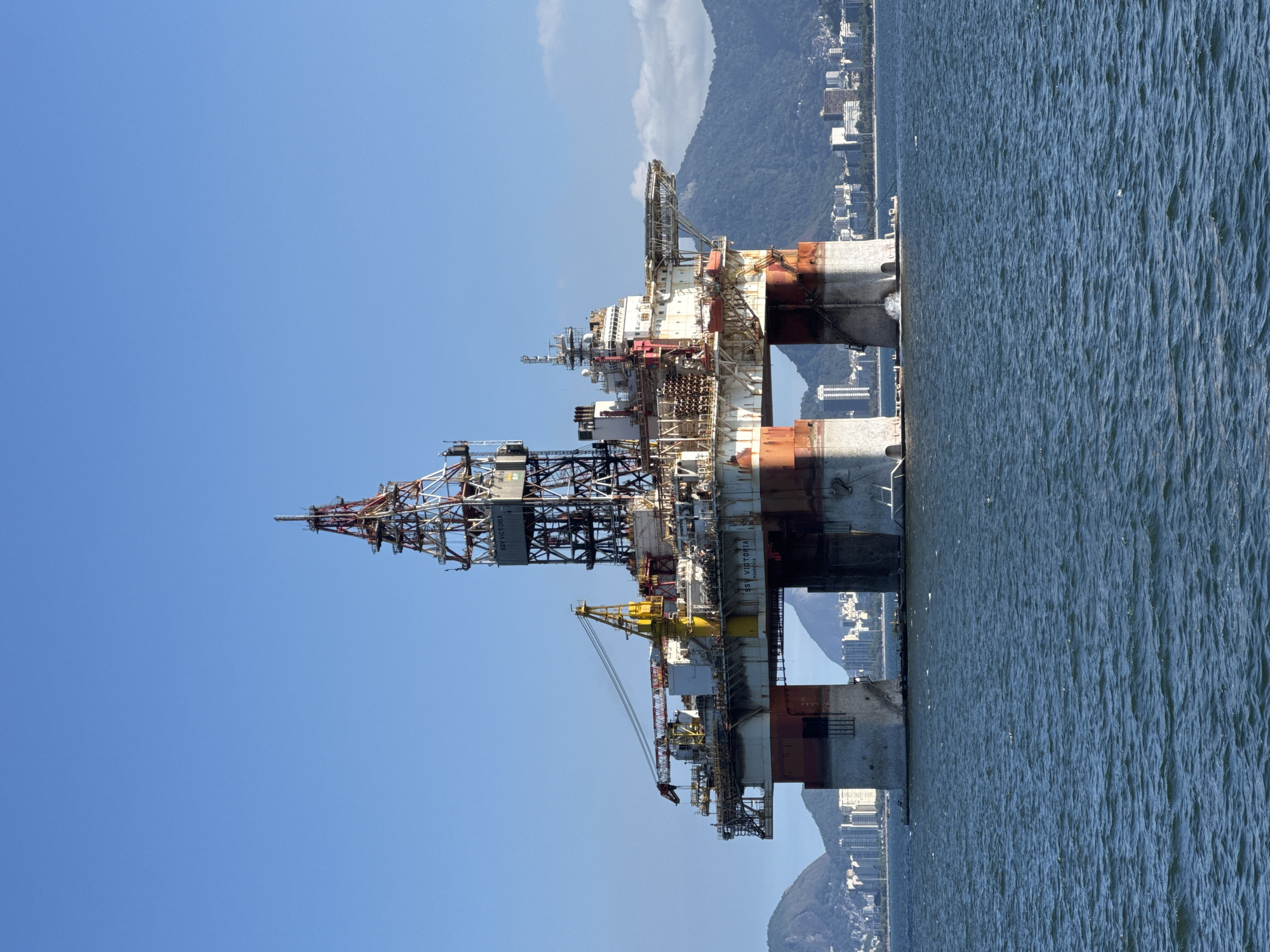 An offshore oil rig named SSV VICTORIA is positioned in the Guanabara Bay, off the coast of Niterói, Brazil. The rig, a semi-submersible platform, is anchored and appears to be operational. It is a large industrial structure with multiple levels, cranes, and a prominent drilling derrick. The platform's lower hull sections are submerged, supporting the deck above the water. The rig is painted in a combination of white, red, and orange colors, showing some signs of weathering.
In the background, the city of Niterói is visible, characterized by its urban skyline with various buildings and structures. Lush green mountains form a backdrop to the city. The water of Guanabara Bay is a deep blue with moderate waves, reflecting the clear blue sky. The lighting suggests it is daytime, with the sun illuminating the scene. No people are discernible on the rig or in the surrounding waters. The overall scene is one of industrial activity juxtaposed with a coastal urban environment and natural landscape.